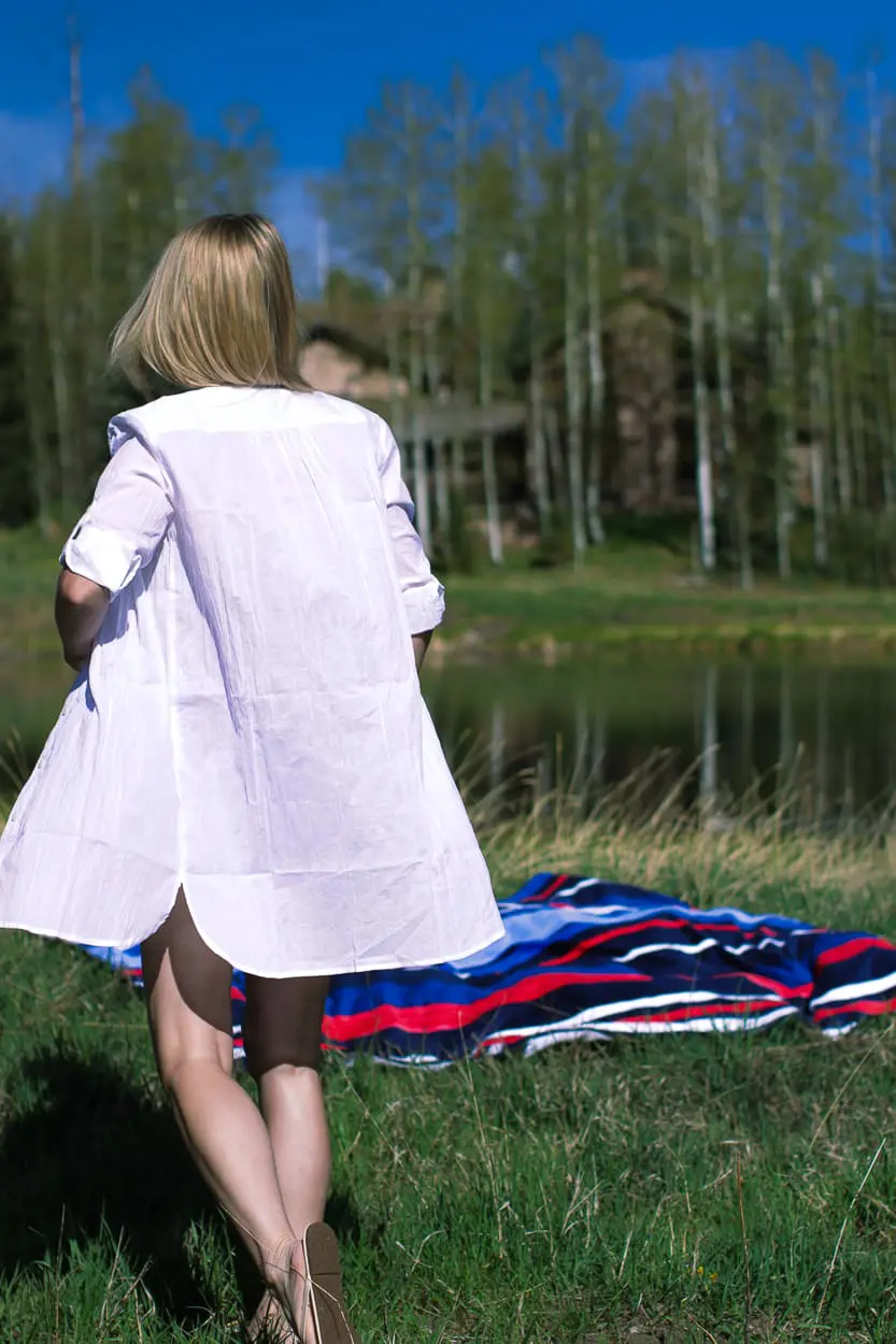 stylish swim shirts and skirts on blonde woman at lake in the mountains wearing a bright blue swim skirt, white button down crinkled cotton swim cover up and a canvas blue and white tote