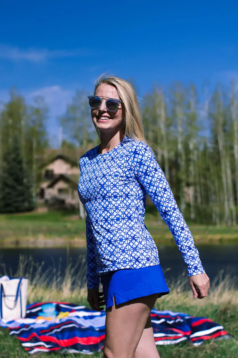 stylish swim shirts and skirts on blonde woman at lake in the mountains wearing a bright blue swim skirt, blue and white geometric swim shirt, white button down crinkled cotton swim cover up and a canvas blue and white tote