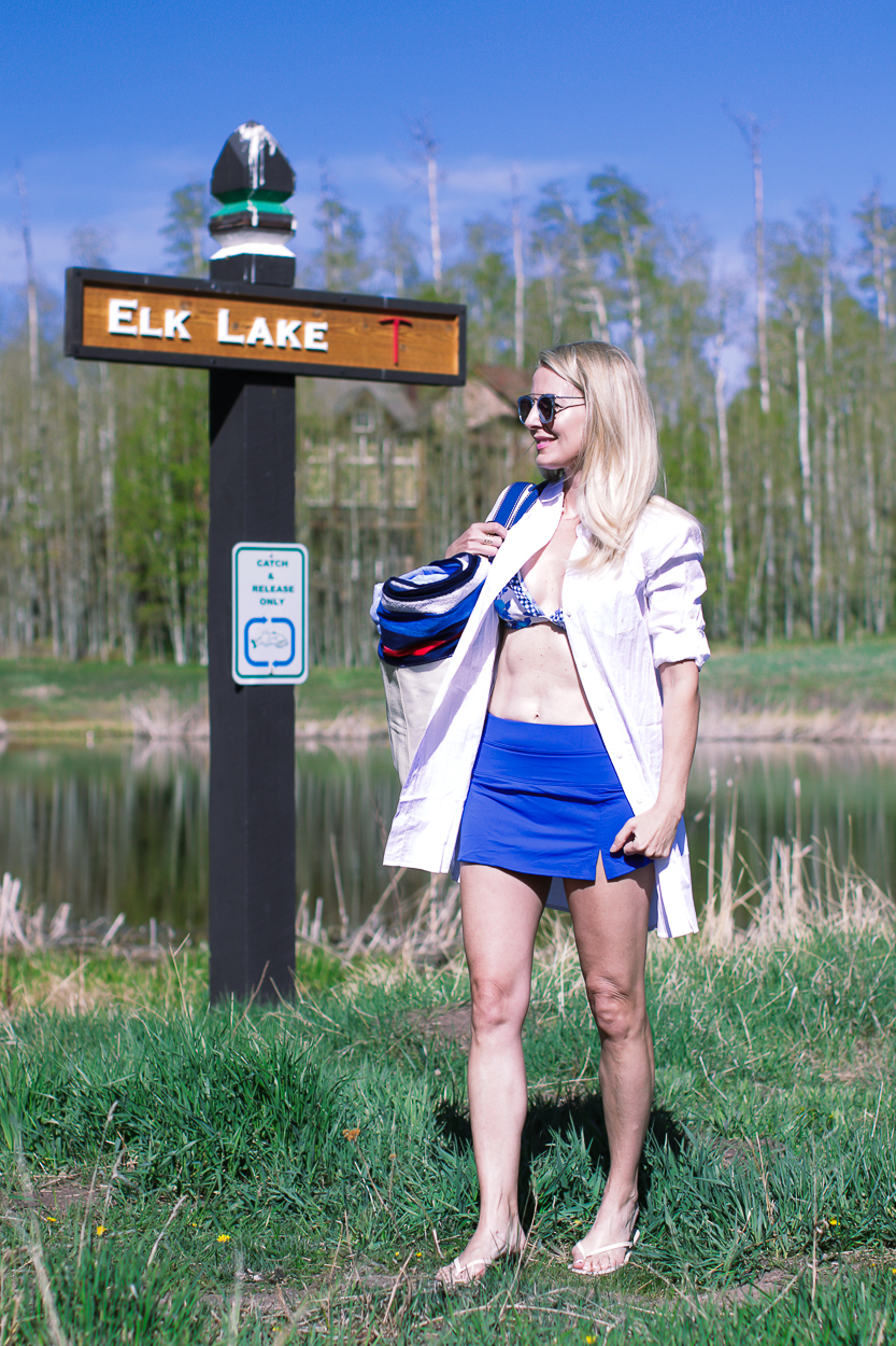 stylish swim shirts and skirts on blonde woman at lake in the mountains wearing a bright blue swim skirt, white button down crinkled cotton swim cover up and a canvas blue and white tote