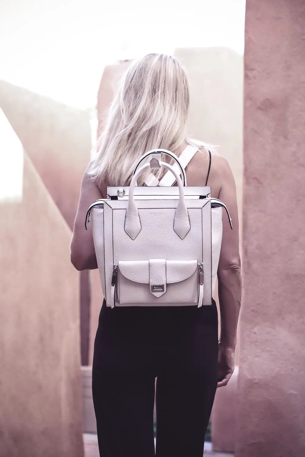 chic and sophisticated backpacks for grown ups by Henri Bendel, featuring blonde woman carrying a white Rivington convertible tote and backpack wearing all black with dainty gold and green jewelry