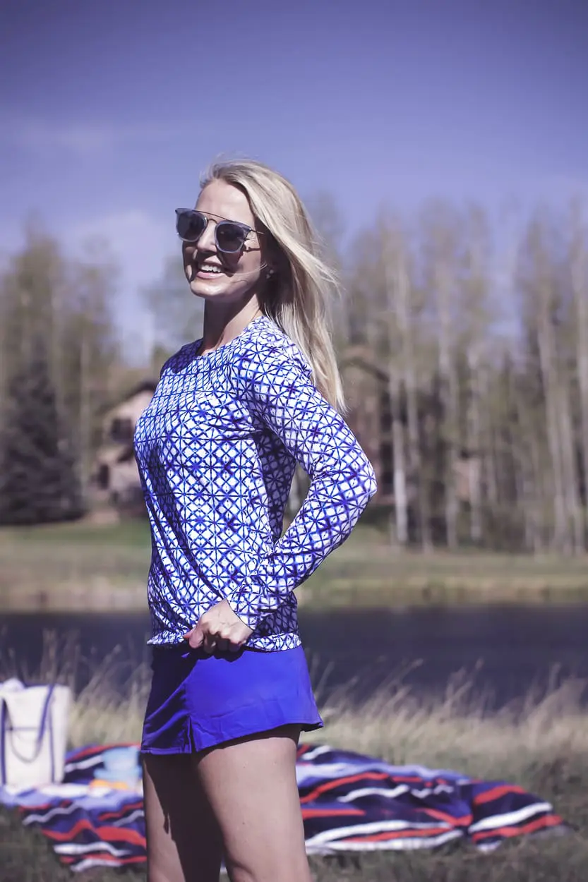 stylish swim shirts and skirts on blonde woman at lake in the mountains wearing a bright blue swim skirt, blue and white geometric swim shirt, white button down crinkled cotton swim cover up and a canvas blue and white tote