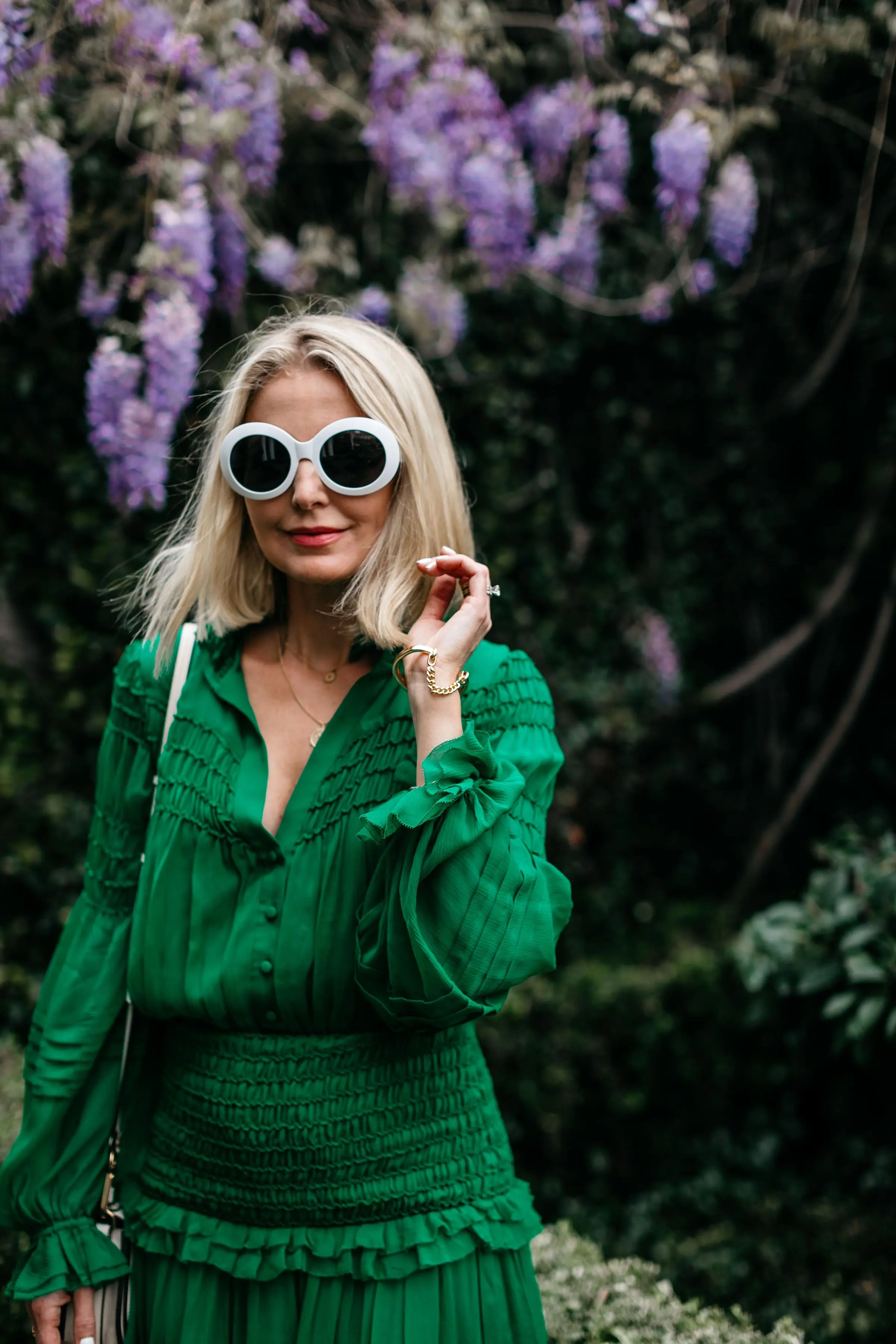 fashion blogger wearing silk green ruffle mini dress posing in front of purple freesia flowers wearing 90s inspired white sunglasses