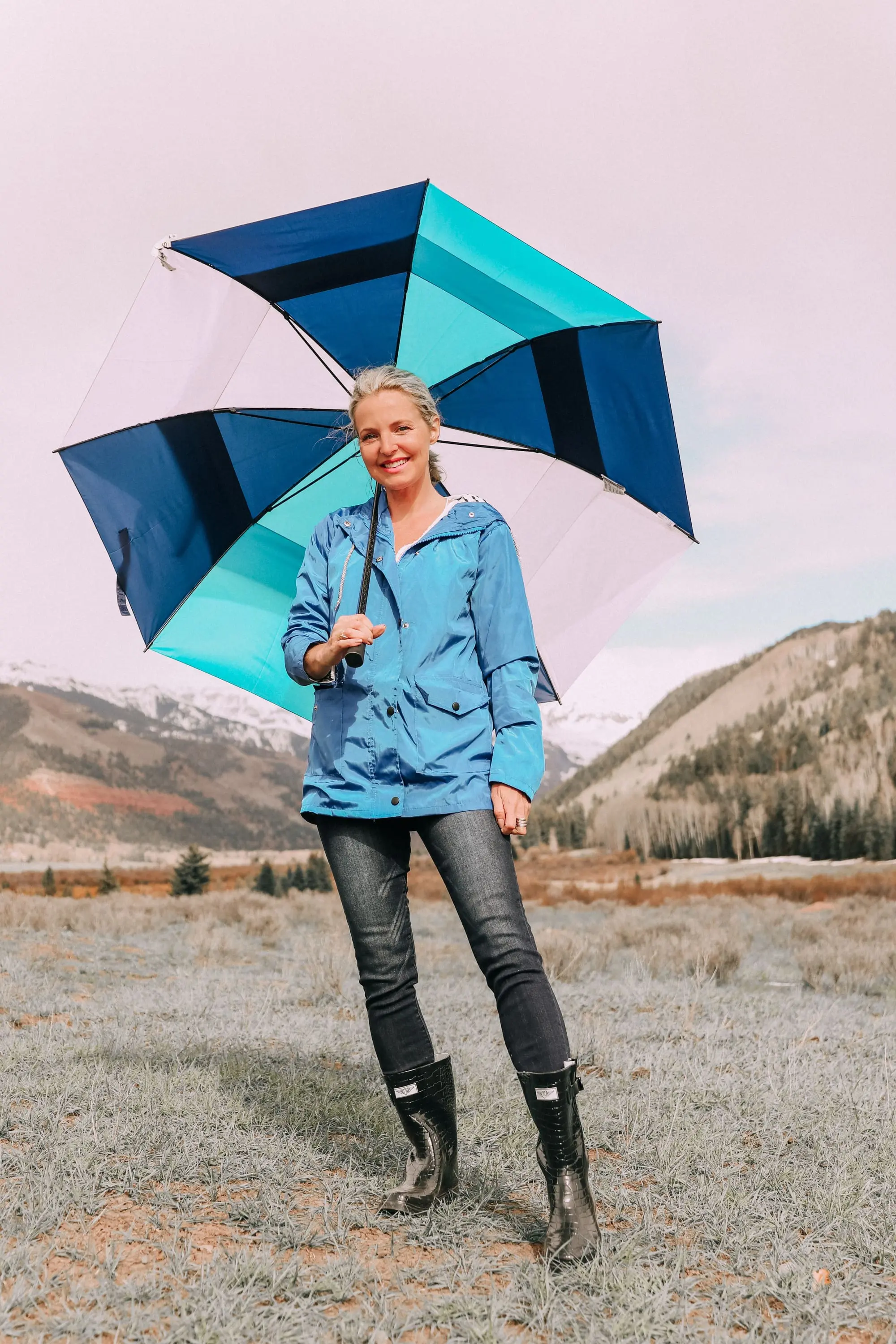 Rainy Day Outfit, Fashion blogger Erin Busbee of BusbeeStyle.com wearing a blue rain jacket, croc-embossed boots, dark wash skinny jeans, and white swiss dot blouse and carrying a colorful blue umbrella from Walmart in Telluride, CO