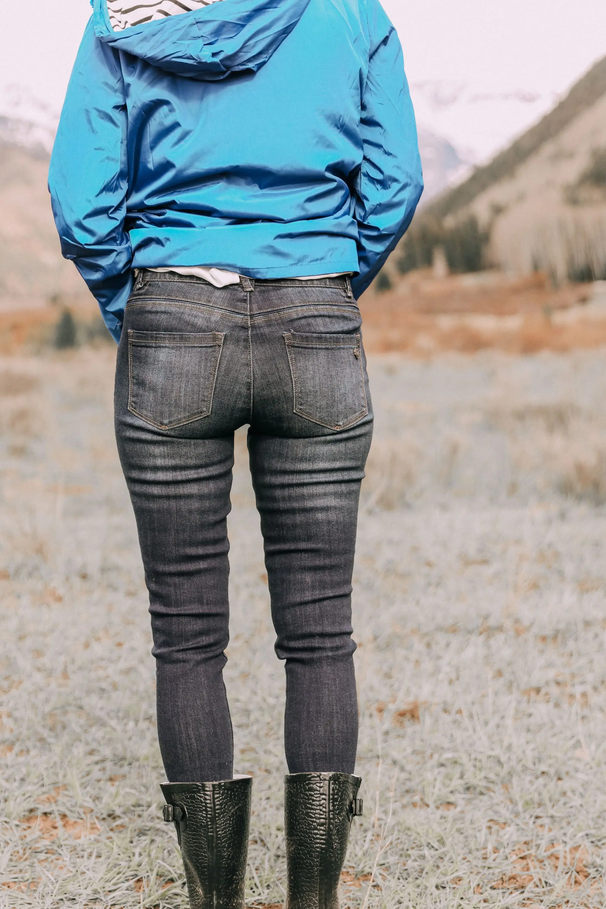 backside of fashion blogger wearing affordable blue waterproof lightweight hooded raincoat paired with dark wash skinny ankle jeans styled with croc patterned rubber rain boots