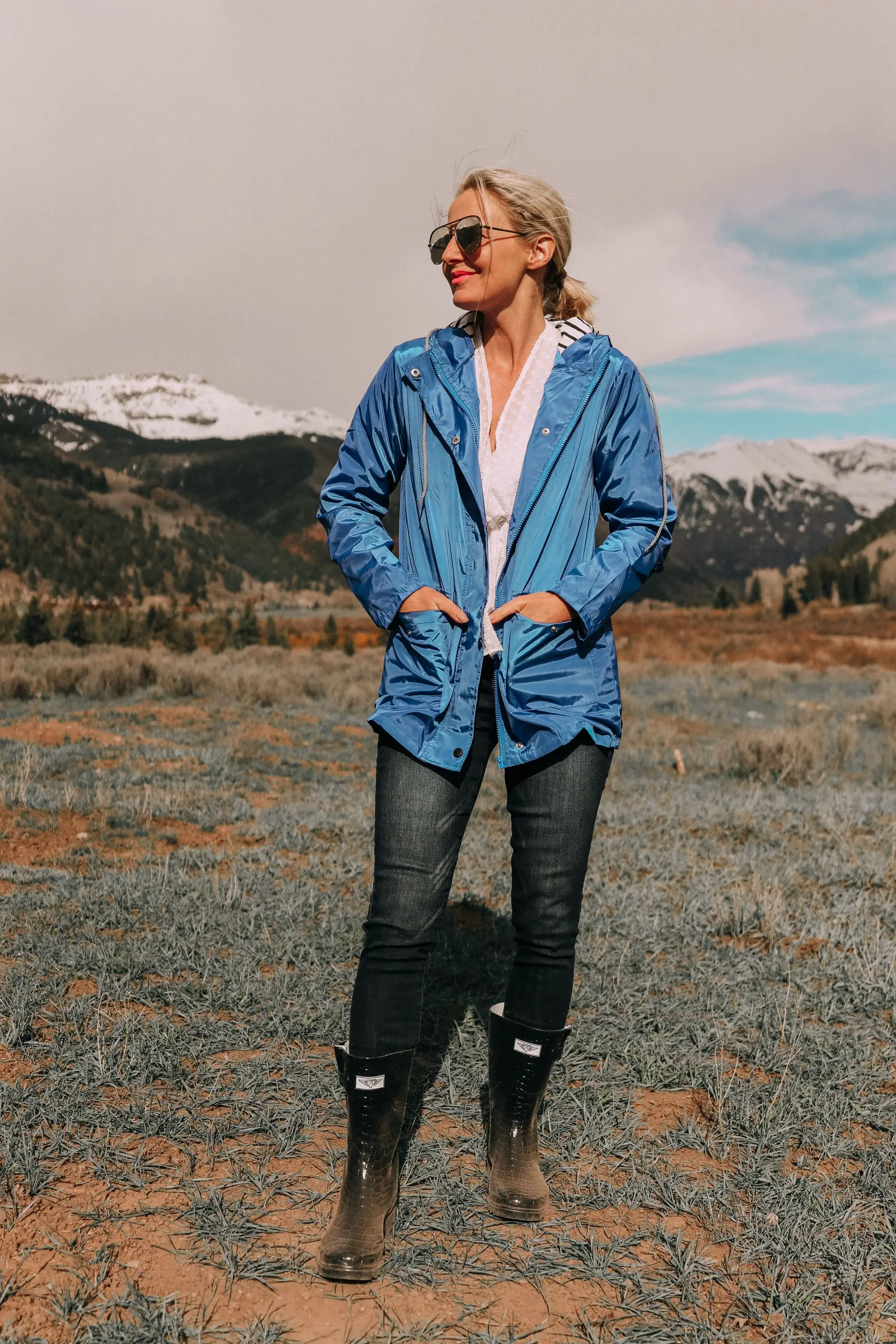 fashion blogger erin busbee in telluride colorado wearing affordable blue waterproof lightweight hooded raincoat over white cinched blouse paired with dark wash skinny ankle jeans styled with croc patterned rubber rain boots
