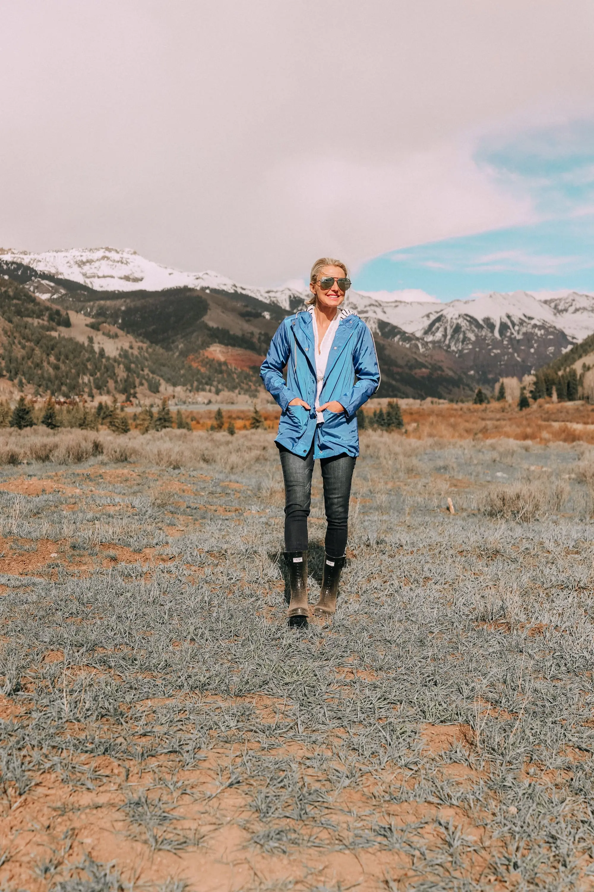 fashion blogger erin busbee in telluride colorado wearing affordable blue waterproof lightweight hooded raincoat over white cinched blouse paired with dark wash skinny ankle jeans styled with croc patterned rubber rain boots