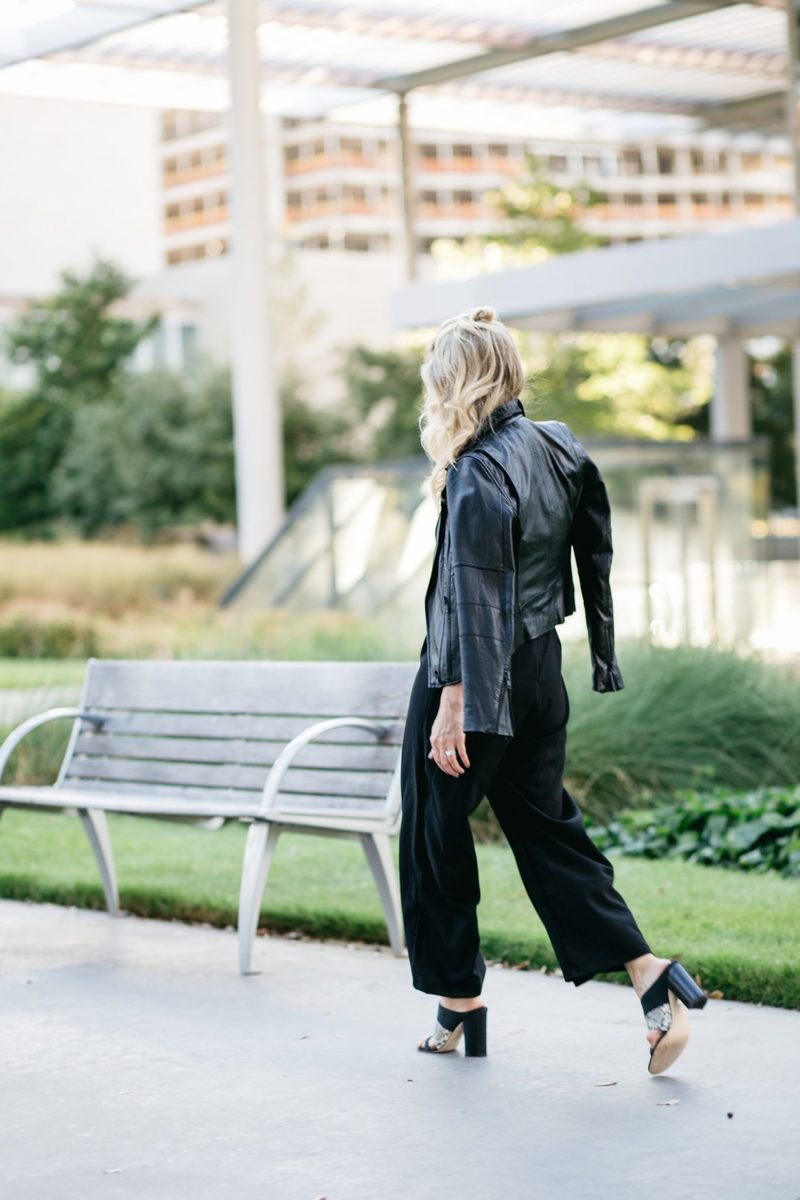 walking away in a black jumpsuit, black moto jacket and slides by m.Gemi