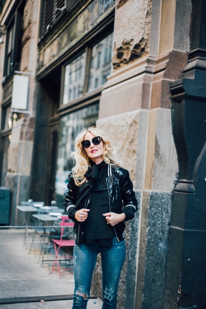 erin busbee in new york city wearing a tie neck black blouse a sequin bomber jacket, skinny, distressed jeans by citizens of humanity with sam edelman mid-calf booties