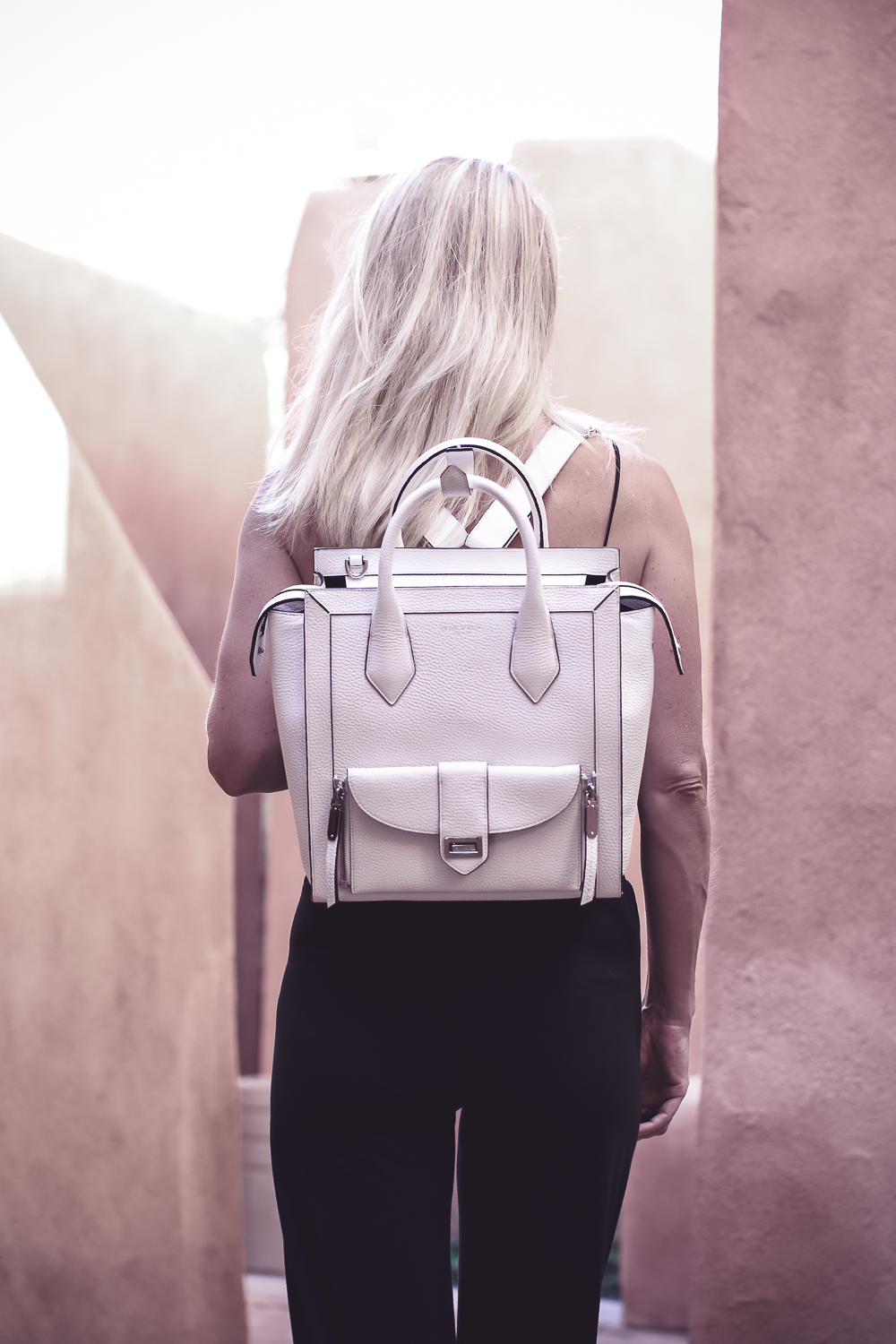 chic and sophisticated backpacks for grown ups by Henri Bendel, featuring blonde woman carrying a white Rivington convertible tote and backpack wearing all black with dainty gold and green jewelry