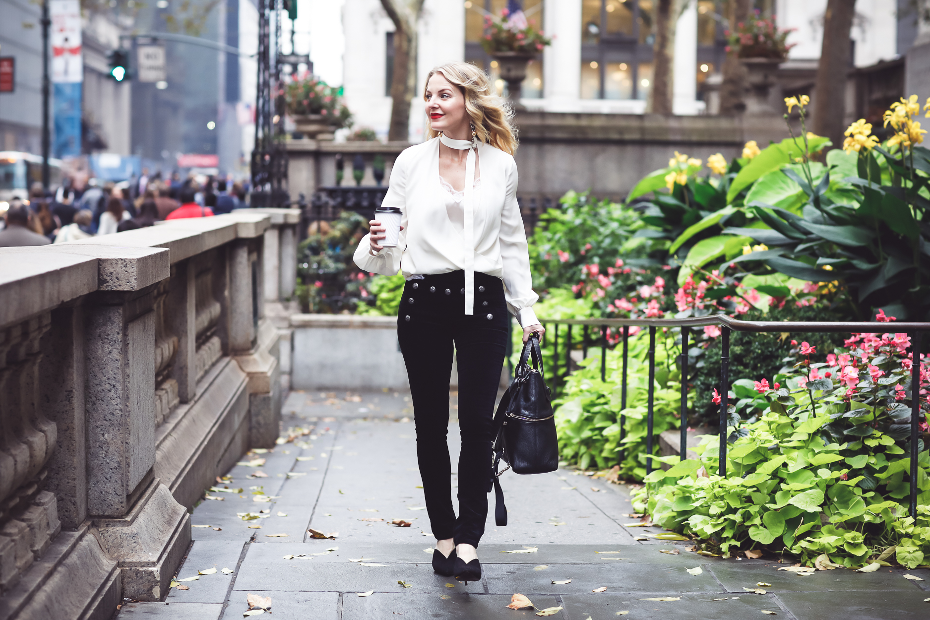 Blonde woman wearing white choker blouse by Alexis, Black velvet veronica beard skinny pants with sailor buttons and black mules by who what wear carrying a Henri Bendel bag in Bryant Park in NYC