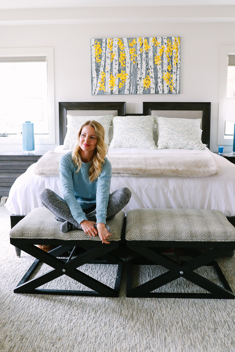 blond woman sitting on bench at foot of king bed with headboard make out of linen and wood, bedding is white and the home is mountain modern