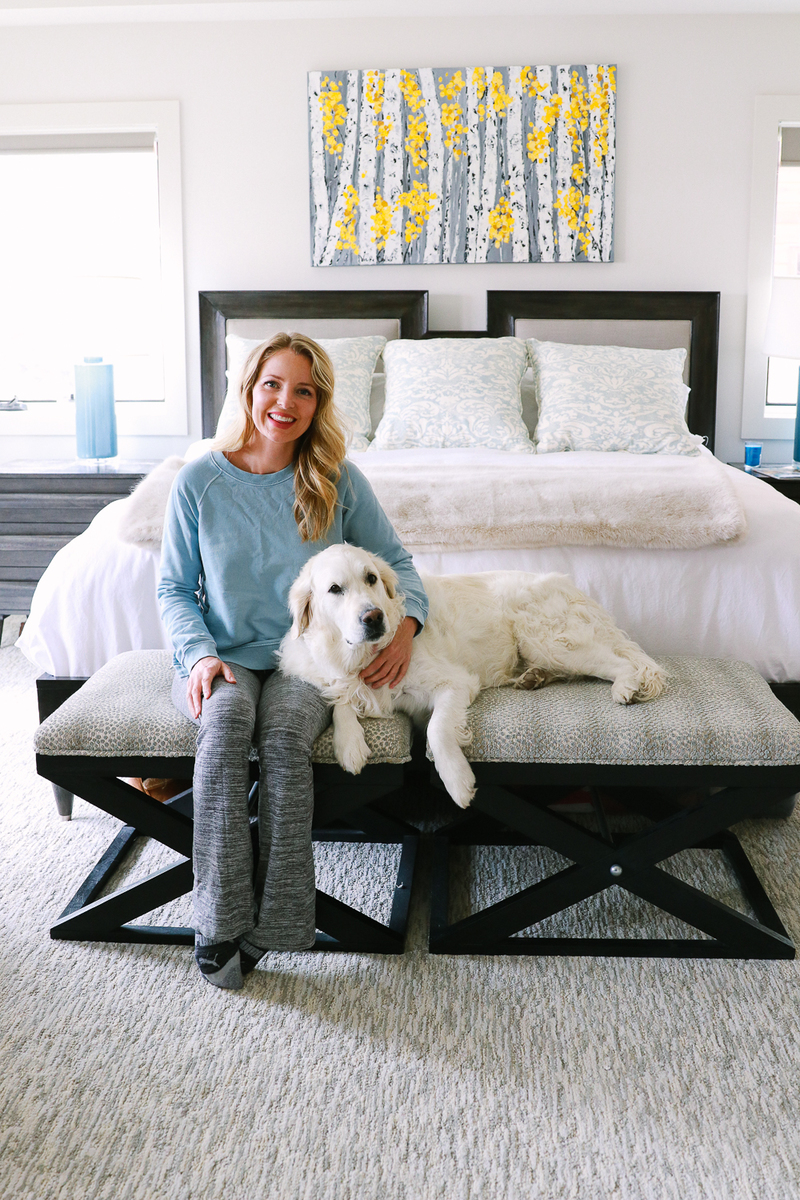 blond woman sitting on bench at foot of king bed with her white golden retriever. the headboard make out of linen and wood, bedding is white and the home is mountain modern