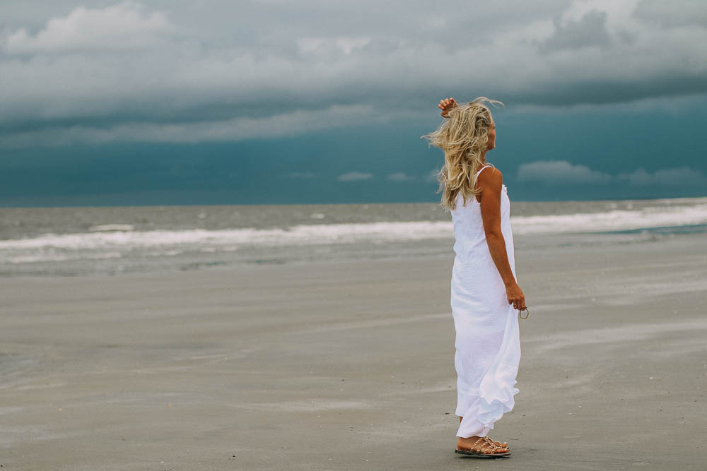Taking blog pictures featuring blurry background, blonde woman on beach wearing white cotton maxi dress