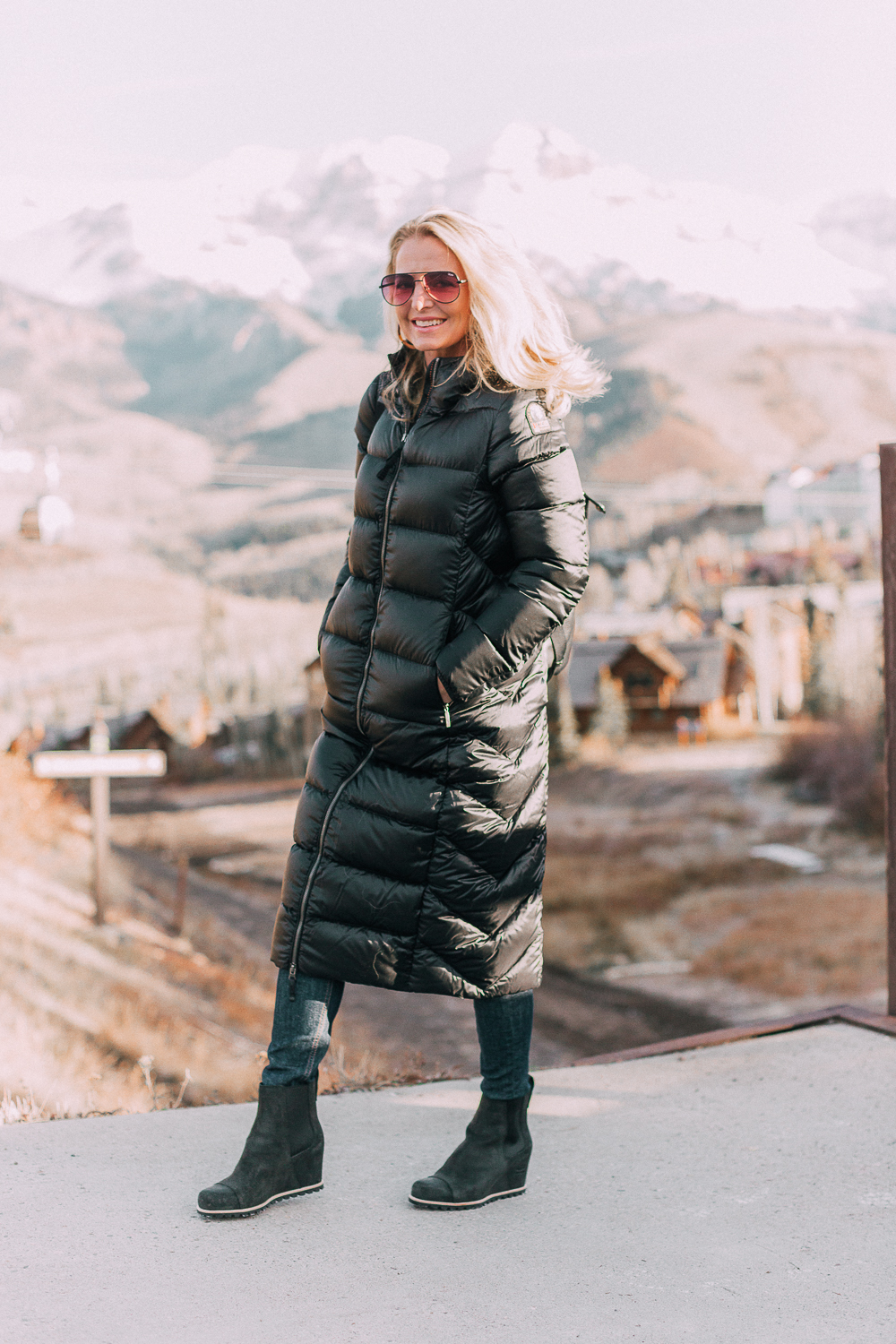 best puffer coats for women by Parajumpers from Bloomingdales in black, long puffer coat paired with dark jeans, black Ugg wedge booties on blonde woman