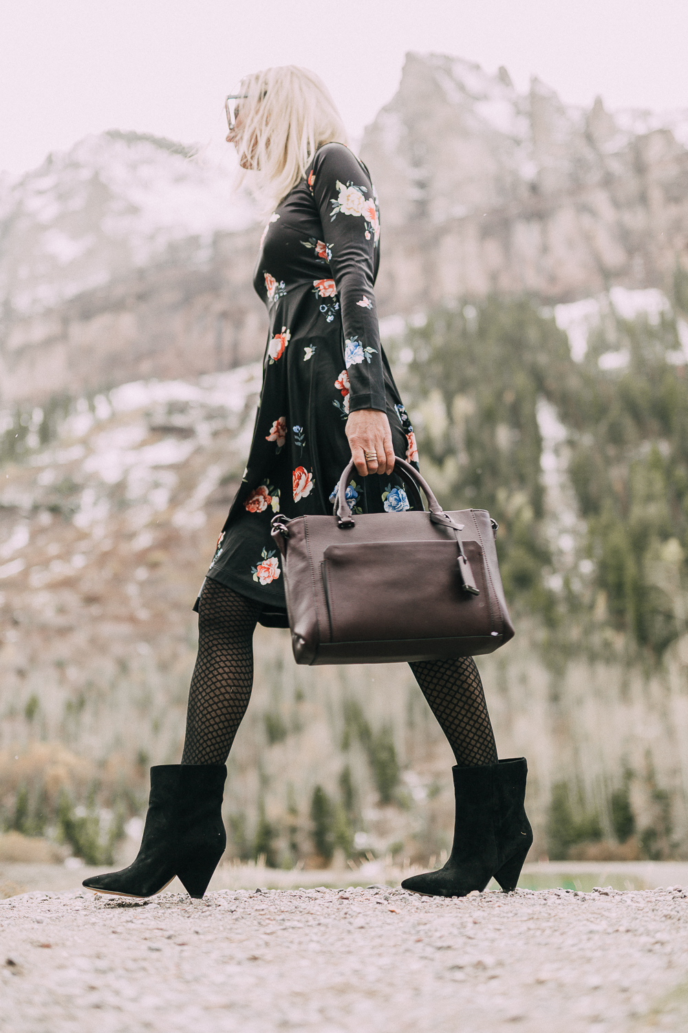 blond woman wearing black vince camuto regina Suede Booties paired with black floral print dress carrying burgundy tote bag with rocky mountains background
