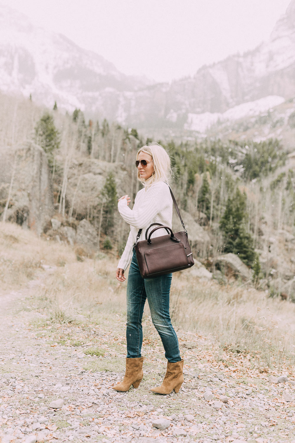 Suede Booties by Vince Camuto paired with white turtleneck cable knit sweater, burgundy tote bag, and white wool coat on blonde woman in the mountains