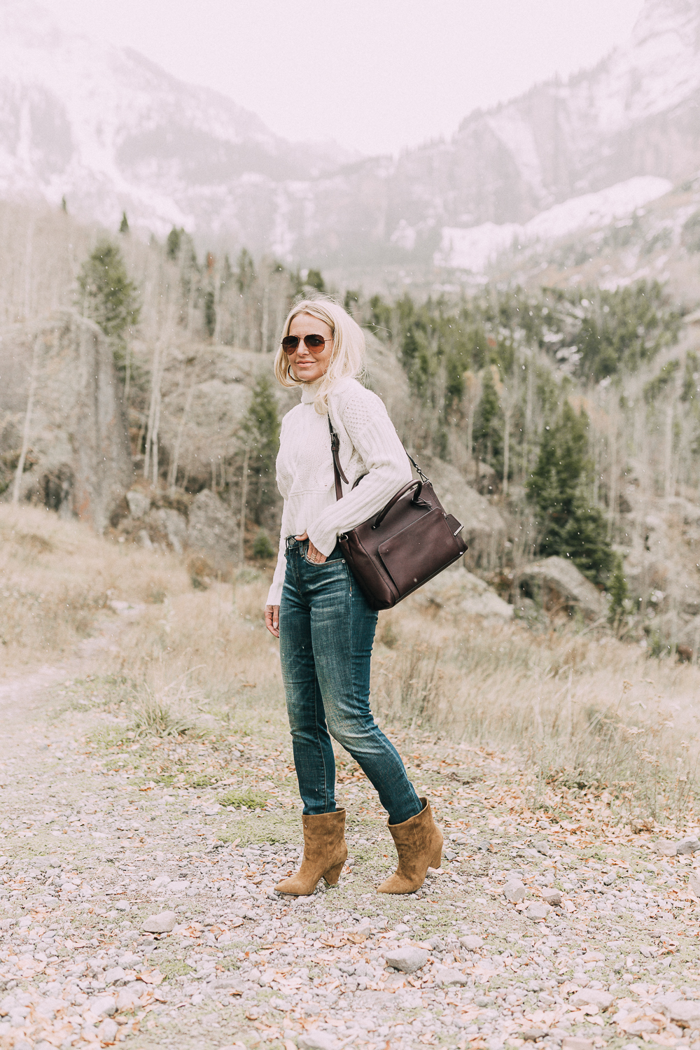 Suede Booties by Vince Camuto paired with white turtleneck cable knit sweater, burgundy tote bag, and white wool coat on blonde woman in the mountains
