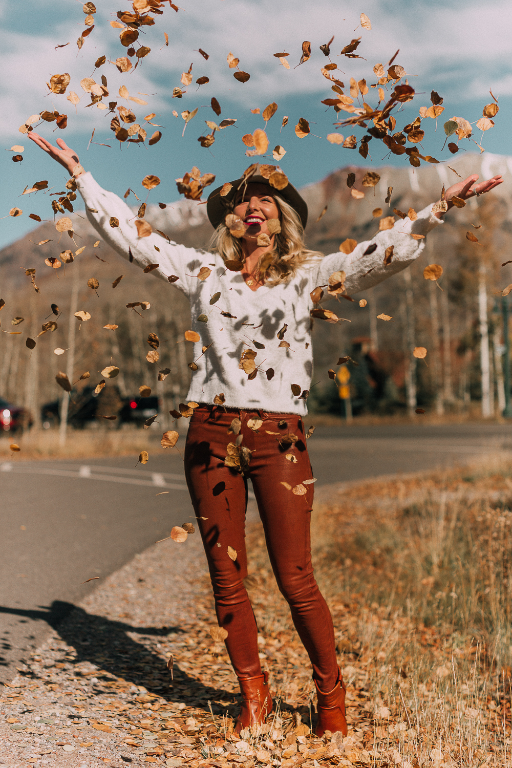 Blonde woman in Telluride, Colorado wearing brown leather pants by Frame with a white fuzzy Halogen sweater, Saint Laurent boots, and a rag & bone hat, throwing leaves in the air 