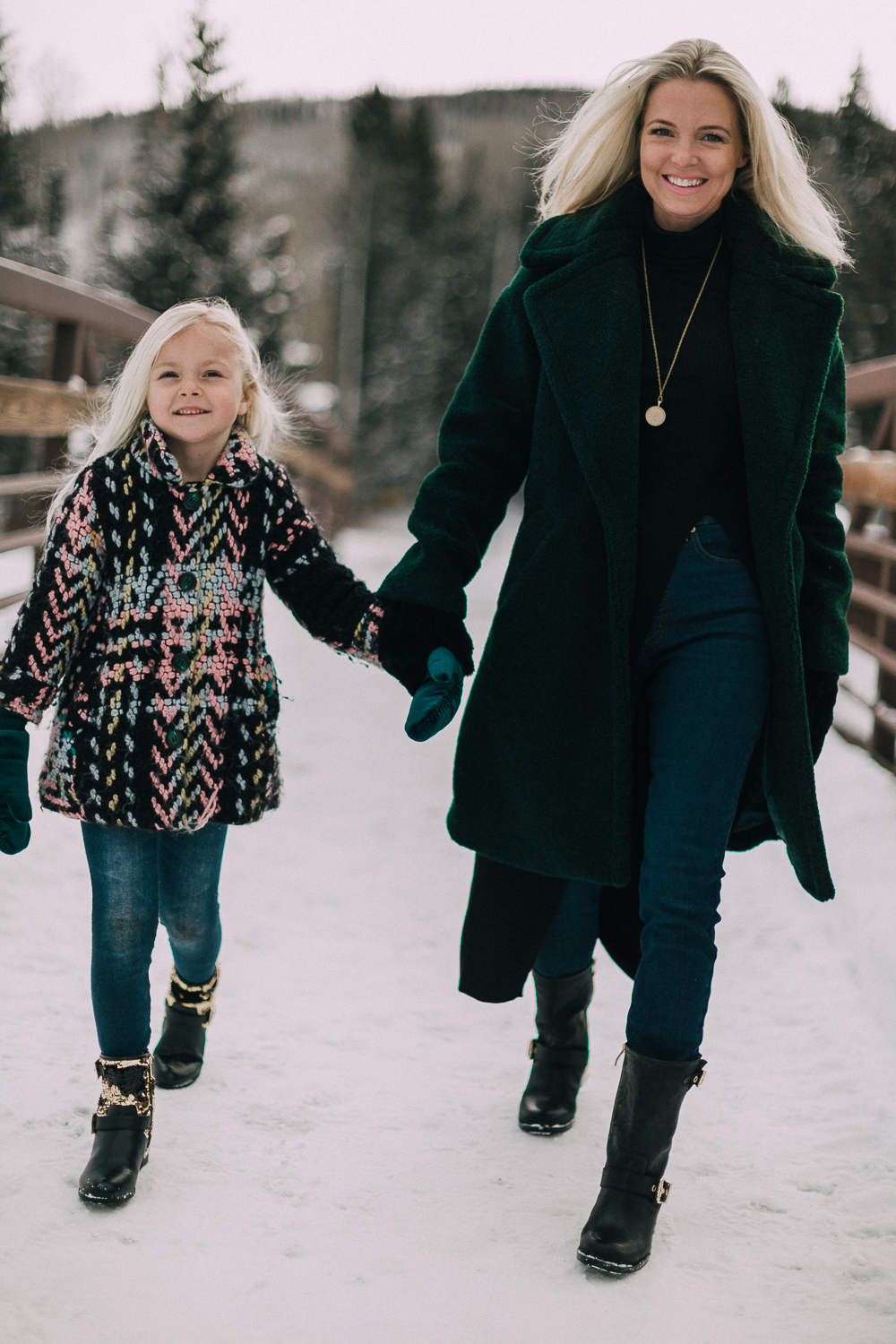 Fashion blogger Erin Busbee of BusbeeStyle.com wearing a green faux fur coat by Kendall & Kylie with black tunic top, jeans, Vince Camuto moto boots, and mittens in Telluride, Colorado walking with her daughter in the snow