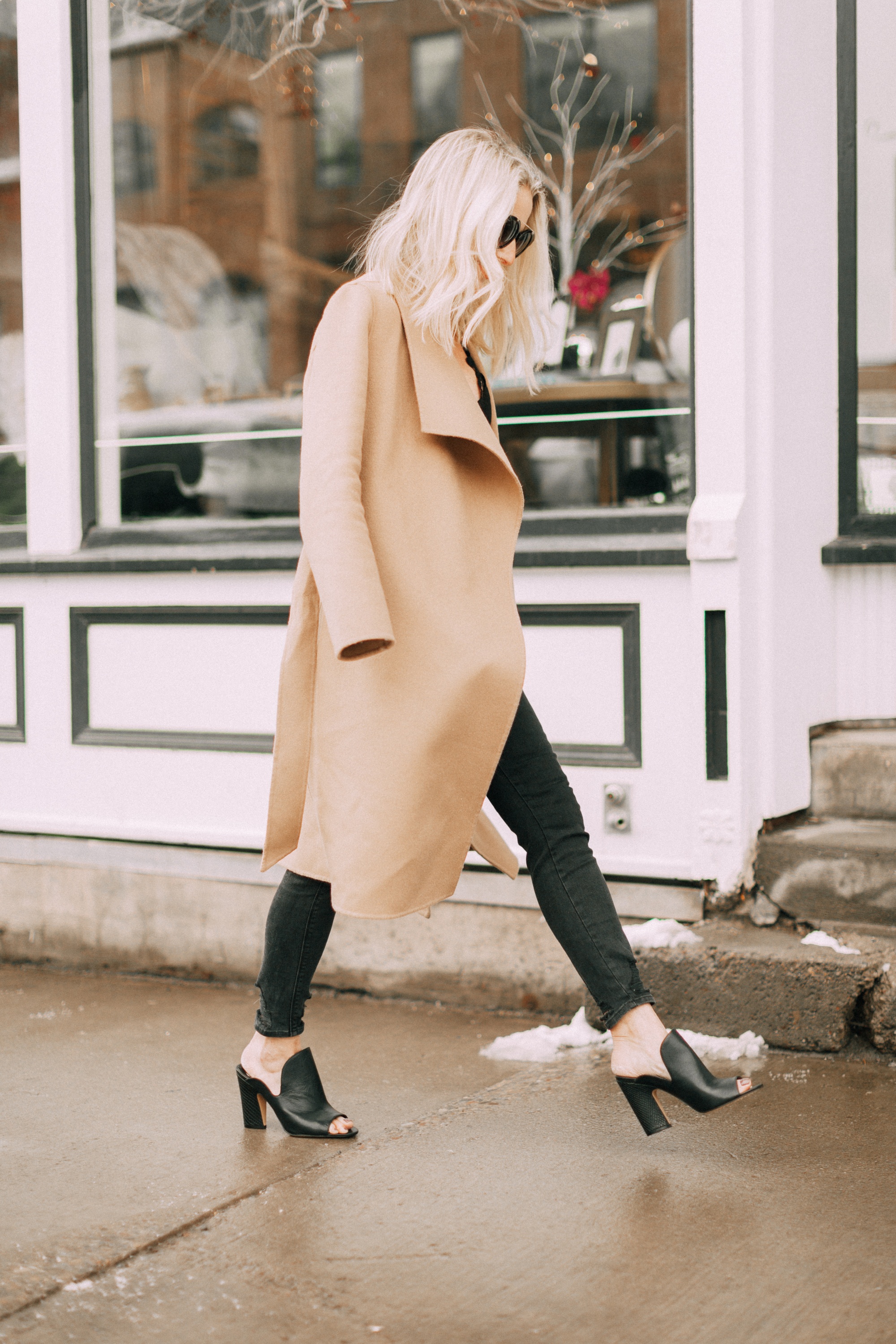blond woman walking on snowy sidewalk telluride colorado wearing long camel coat black j brand jeans black Vince Camuto mules