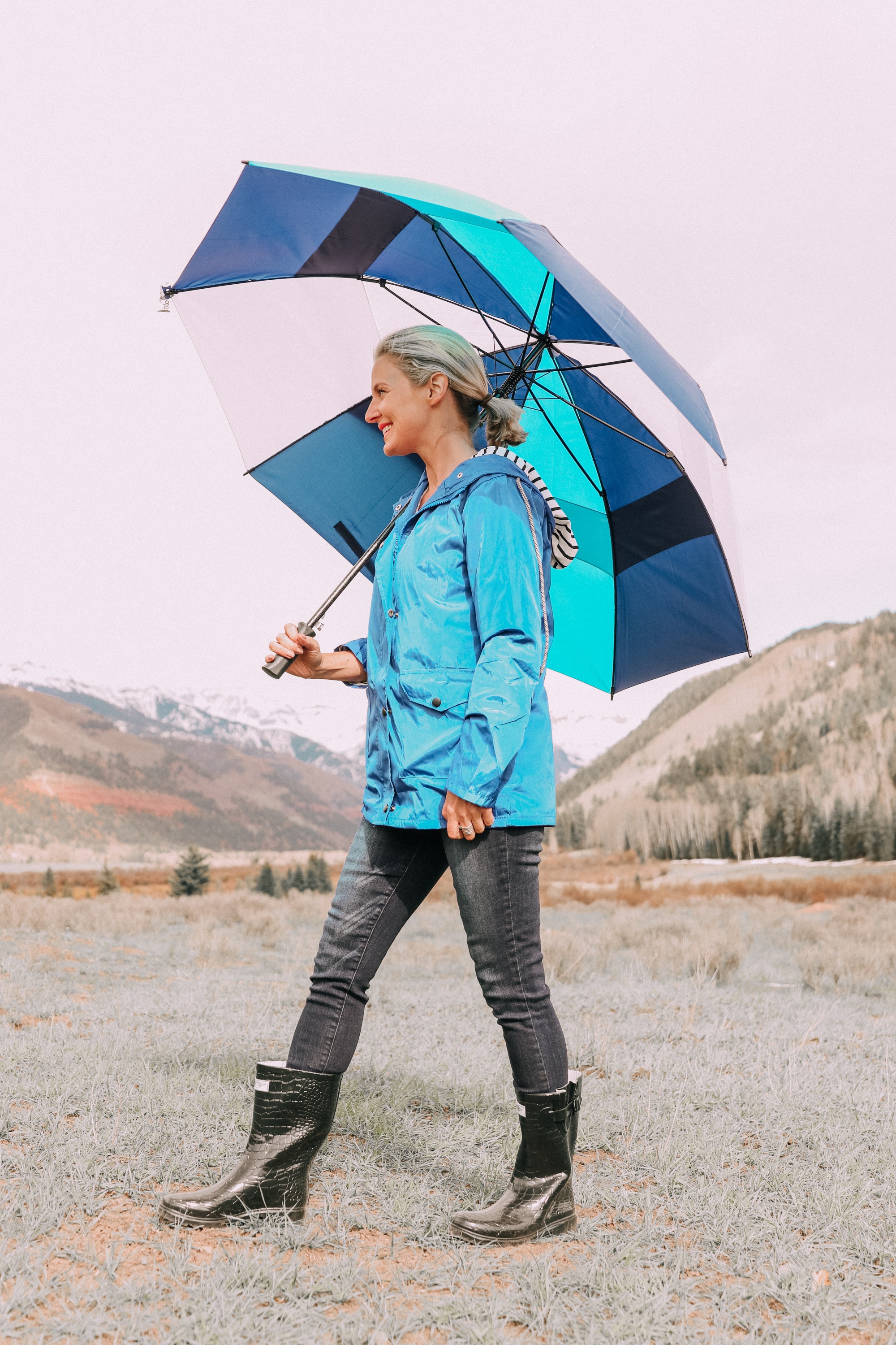 fashion blogger erin busbee in telluride colorado wearing affordable blue waterproof lightweight hooded raincoat paired with dark wash skinny ankle jeans croc patterned rubber rain boots holding blue and white totes umbrella 