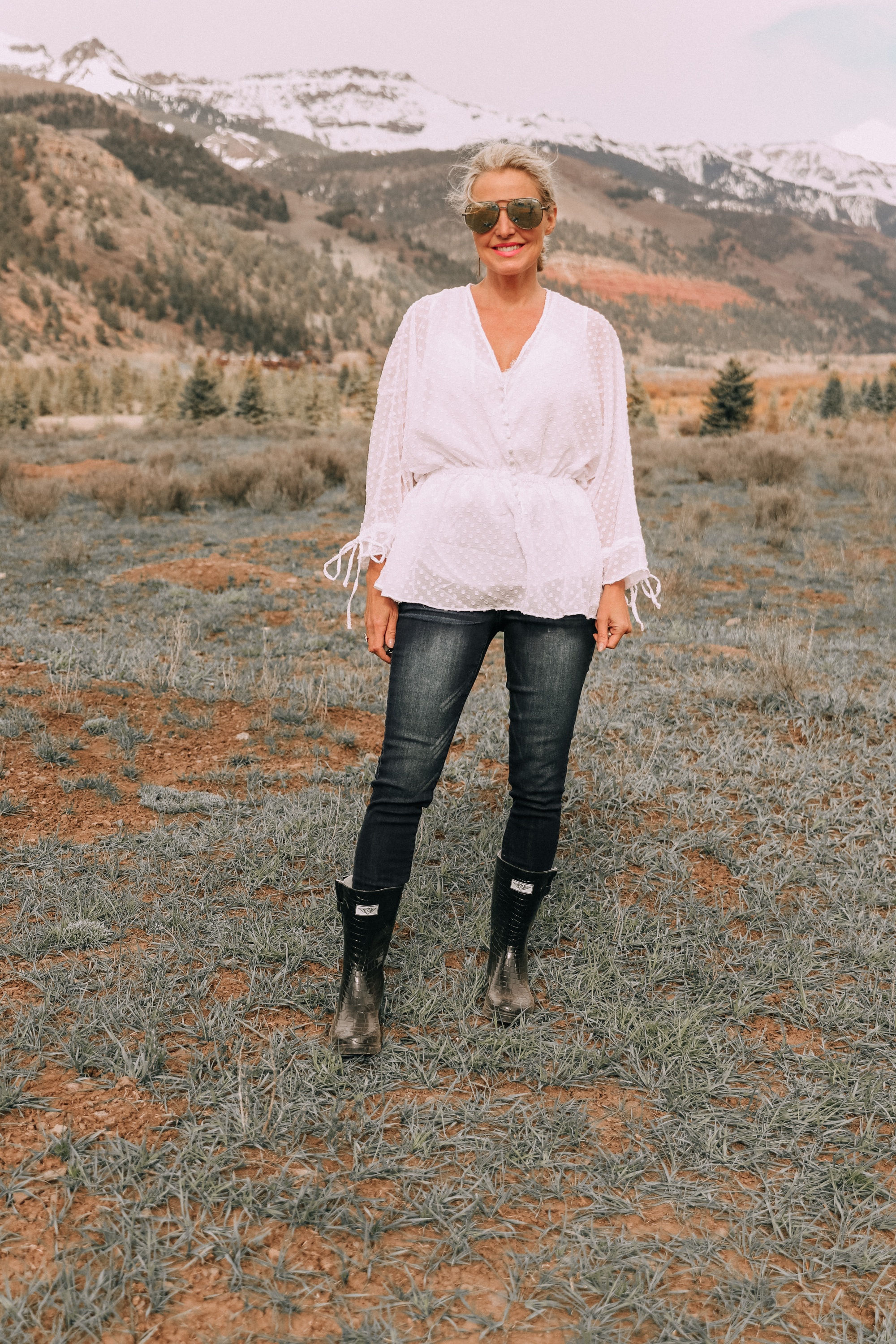 fashion blogger erin busbee in telluride colorado wearing white cinched blouse paired with dark wash skinny ankle jeans styled with croc patterned rubber rain boots