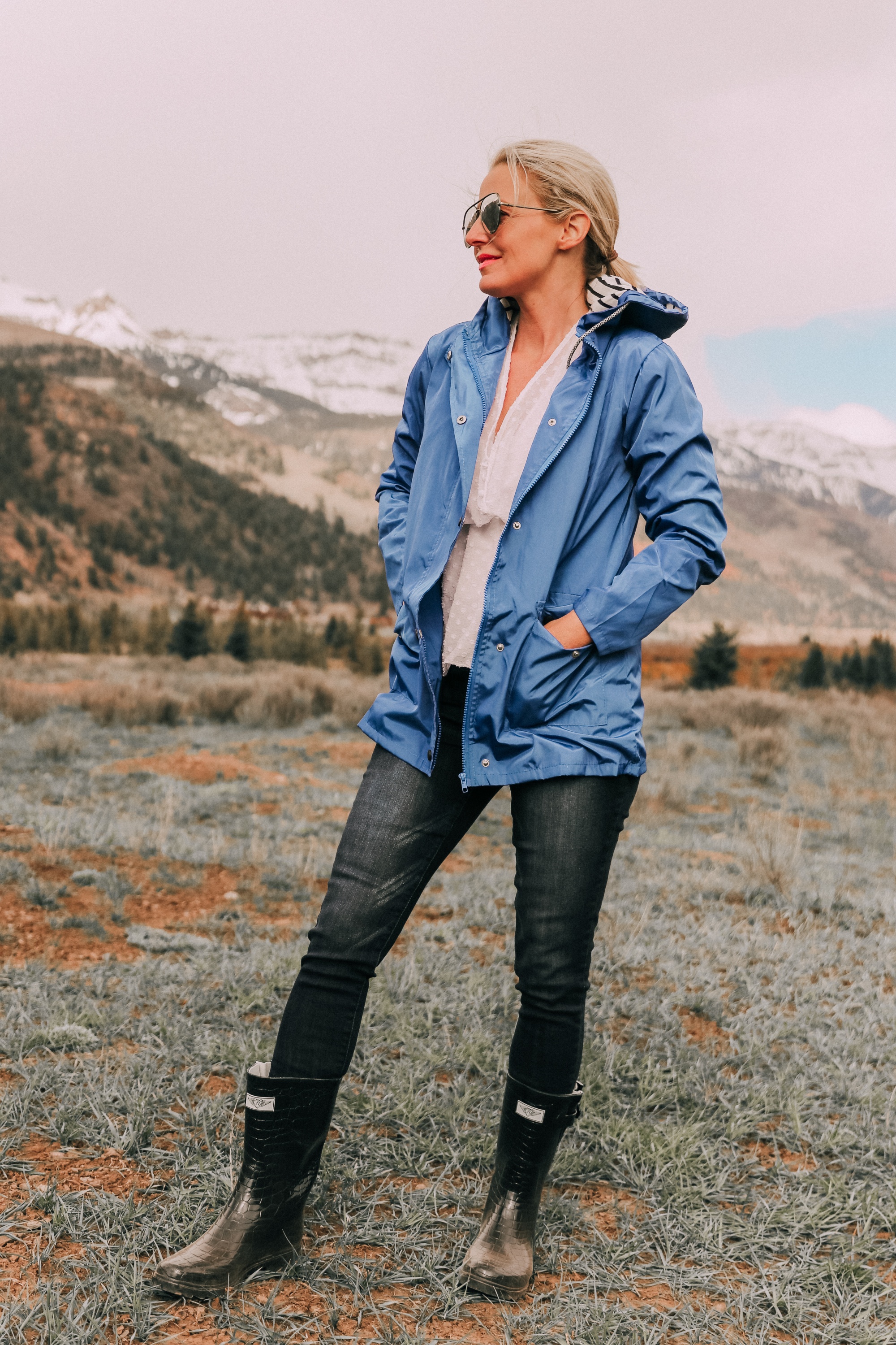 fashion blogger erin busbee in telluride colorado wearing affordable blue waterproof lightweight hooded raincoat over white cinched blouse paired with dark wash skinny ankle jeans styled with croc patterned rubber rain boots