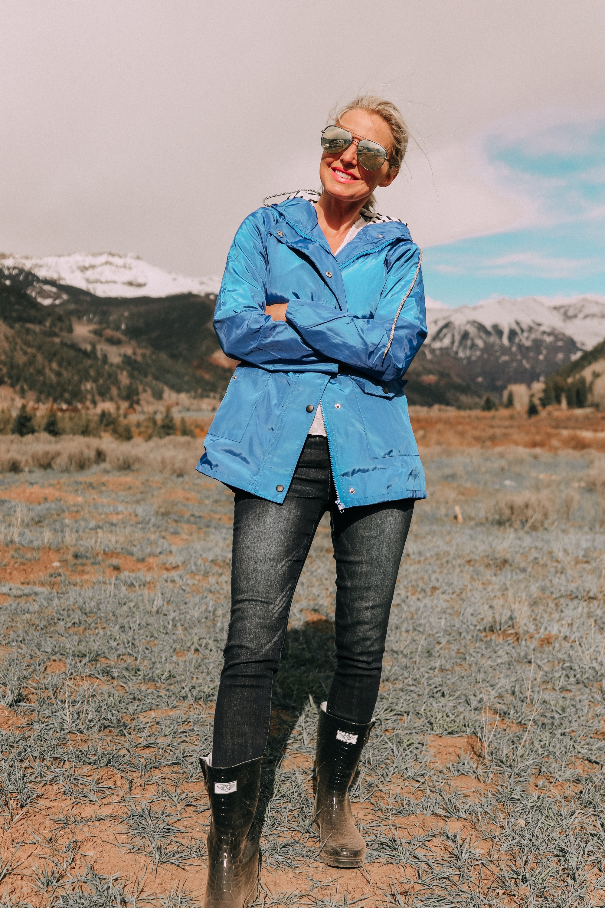 fashion blogger erin busbee in telluride colorado wearing affordable blue waterproof lightweight hooded raincoat over white cinched blouse paired with dark wash skinny ankle jeans styled with croc patterned rubber rain boots