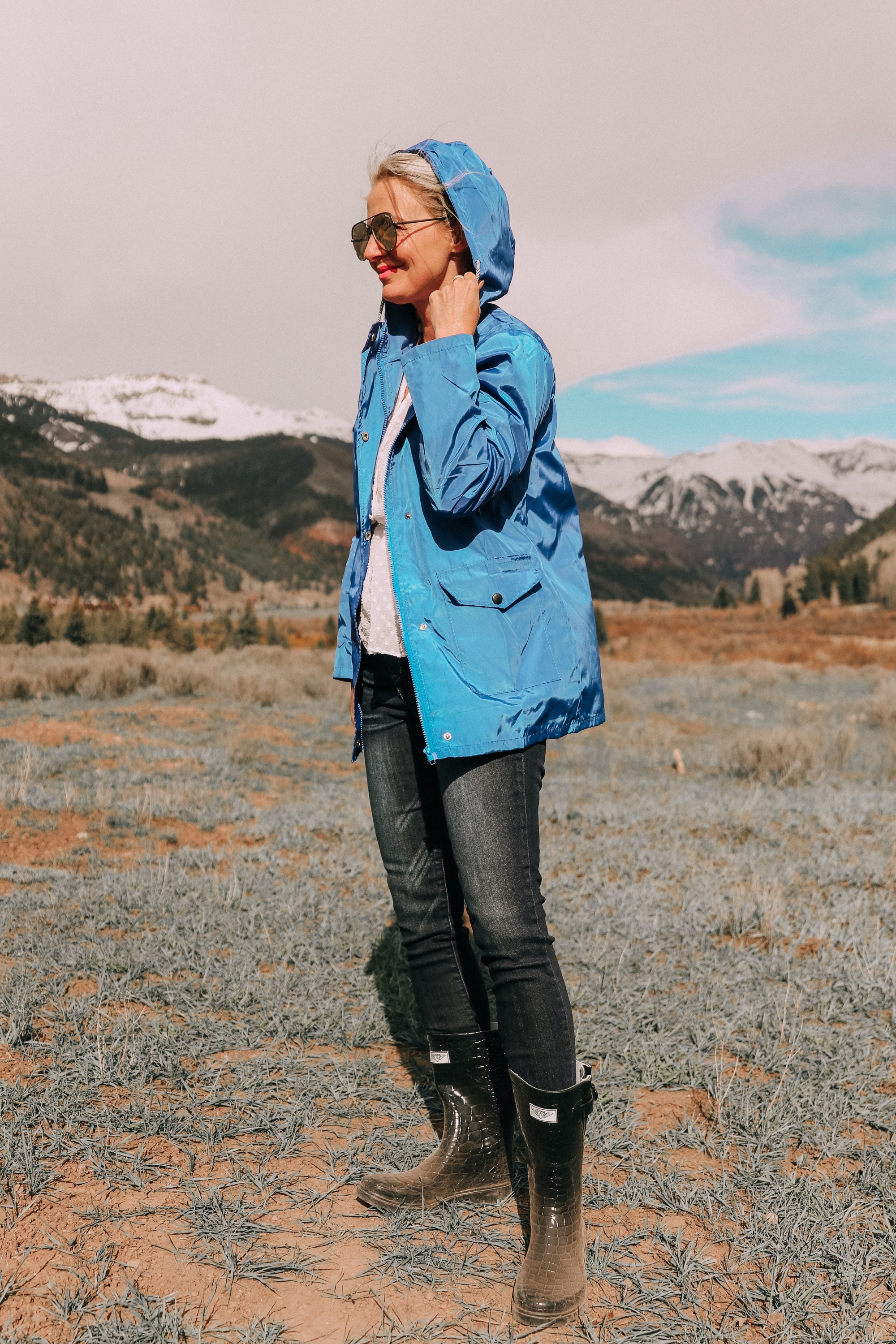 fashion blogger erin busbee in telluride colorado wearing affordable blue waterproof lightweight hooded raincoat with hood over head paired with dark wash skinny ankle jeans styled with croc patterned rubber rain boots