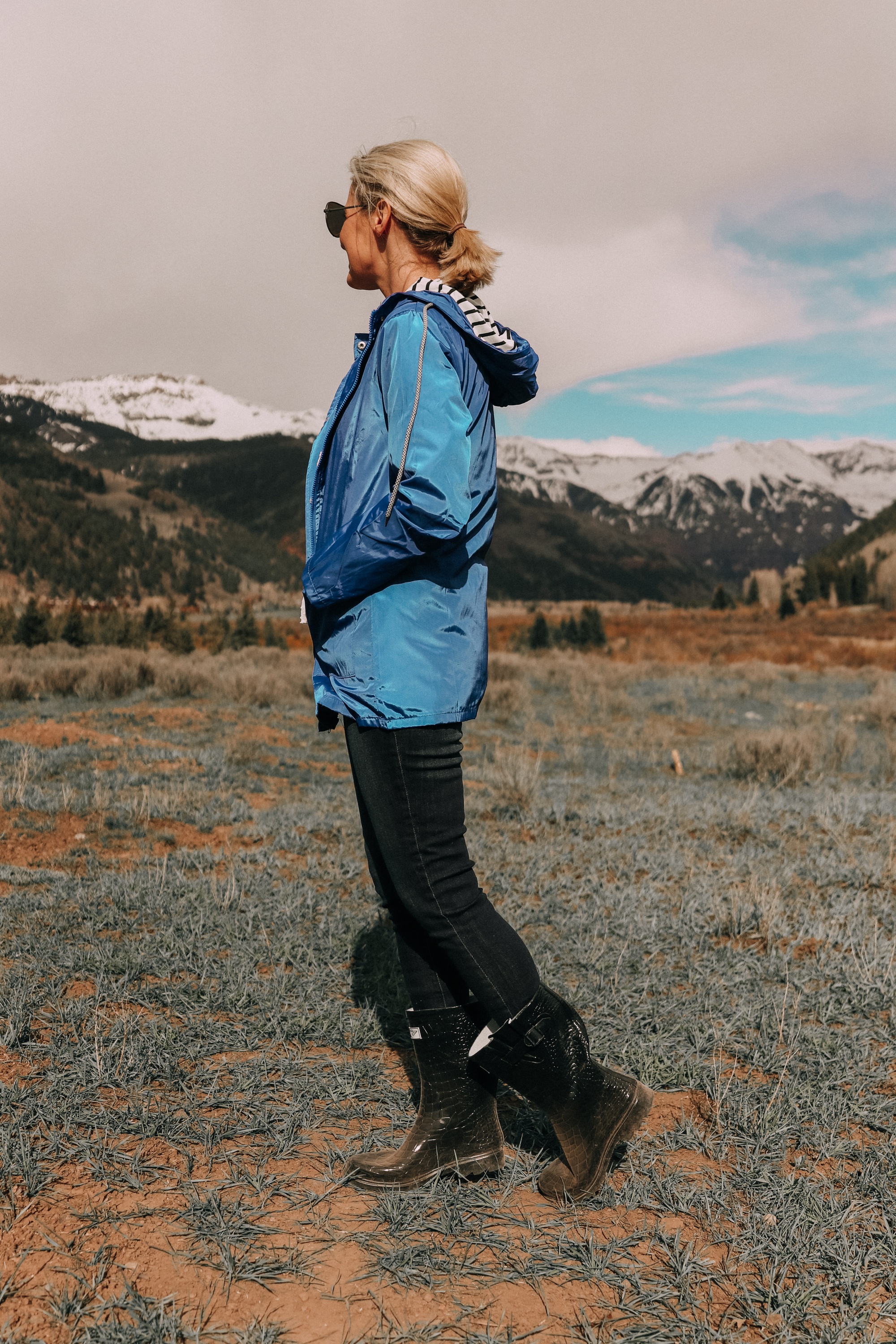 sideview of fashion blogger erin busbee in telluride colorado wearing affordable blue waterproof lightweight striped hooded raincoat with dark wash skinny ankle jeans paired with croc patterned rubber rain boots
