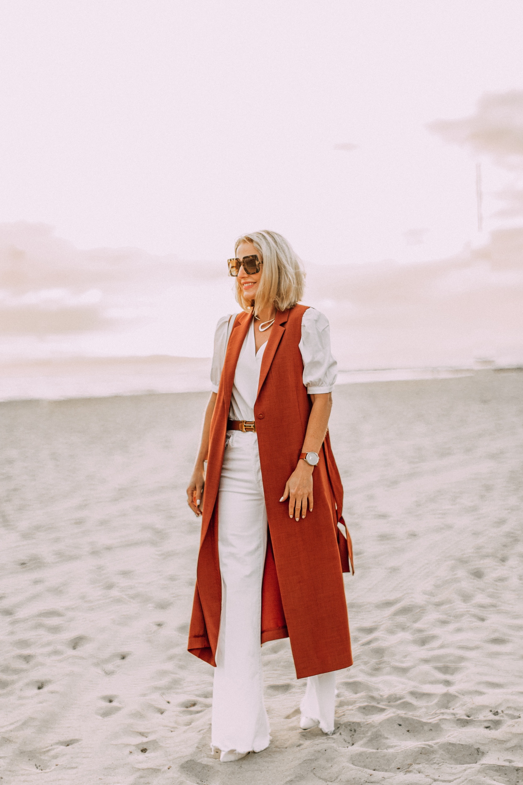 fashion blogger wearing BCBG long rust colored vest over all white outfit wth hermes belt on santa monica beach