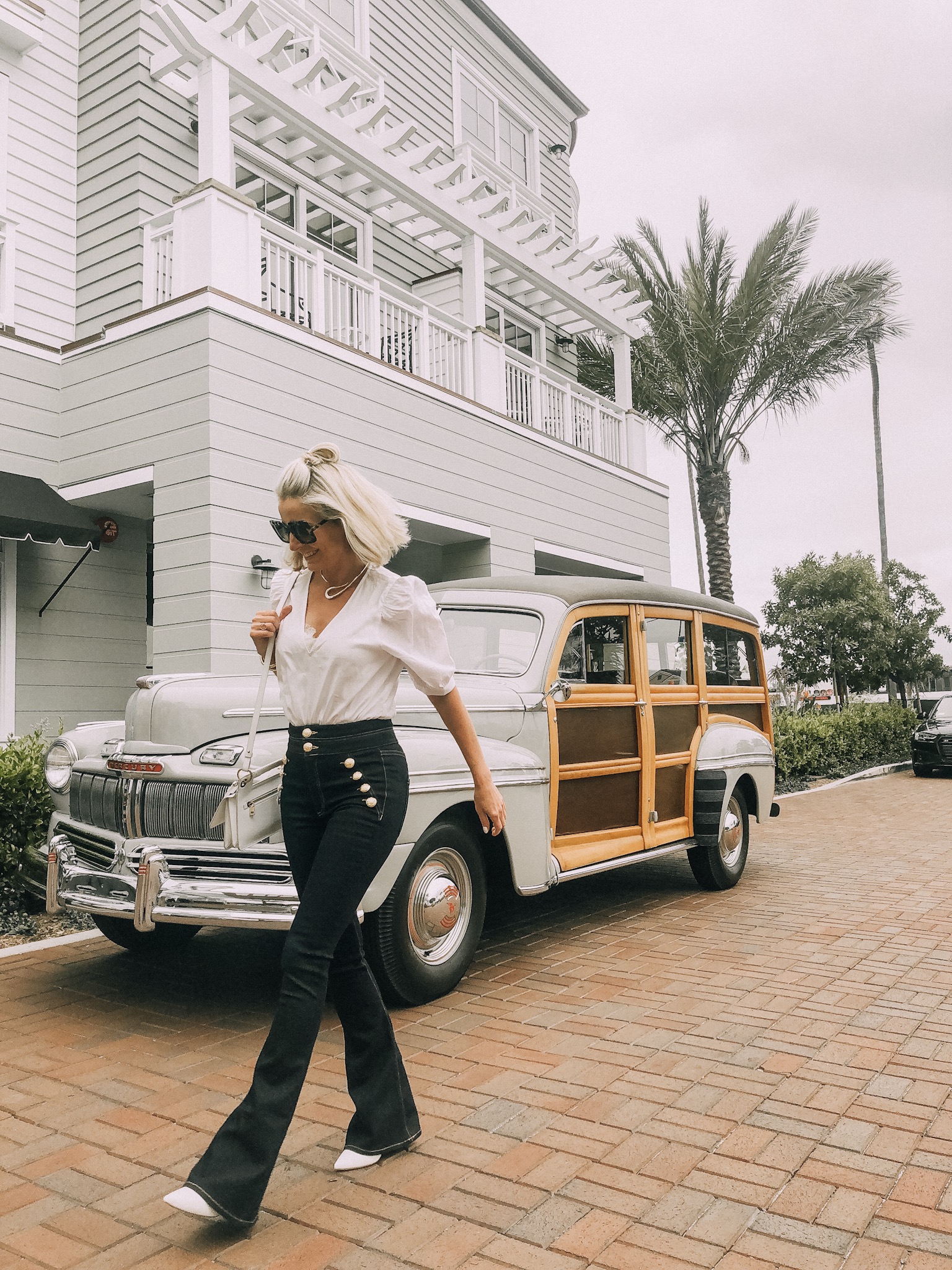 fashion blogger wearing veronica beard blue jeans white bodysuit walking past vintage woody car on newport beach california