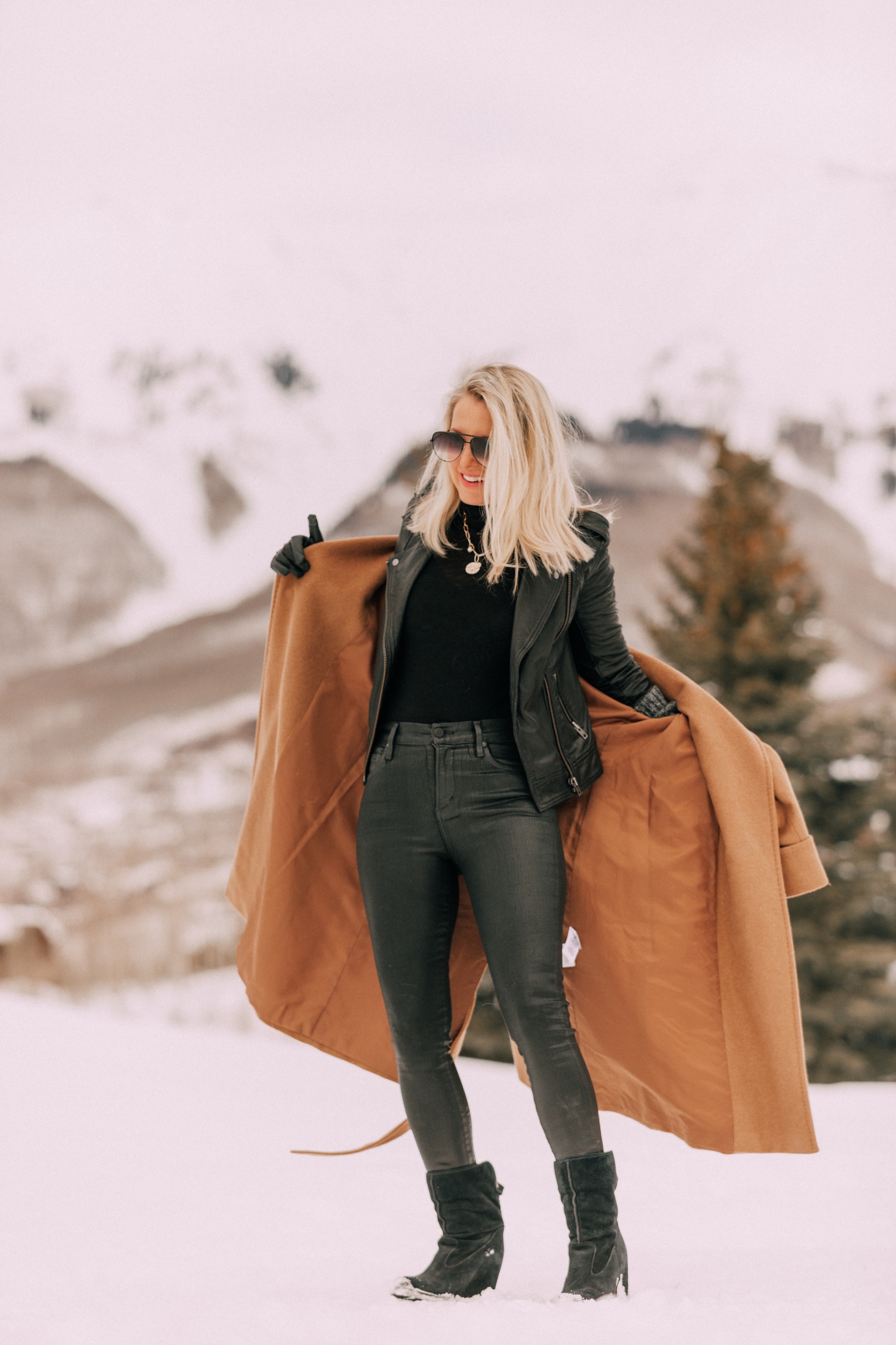 How To Layer A Moto Jacket, Fashion blogger Erin Busbee of BusbeeStyle.com wearing a black leather IRO moto jacket, Carolina Amato gloves, rocket leatherette skinny jeans by Citizens of Humanity, black cashmere turtleneck, and Ugg wedge boots and putting on a wool camel coat by Mackage in Telluride, CO