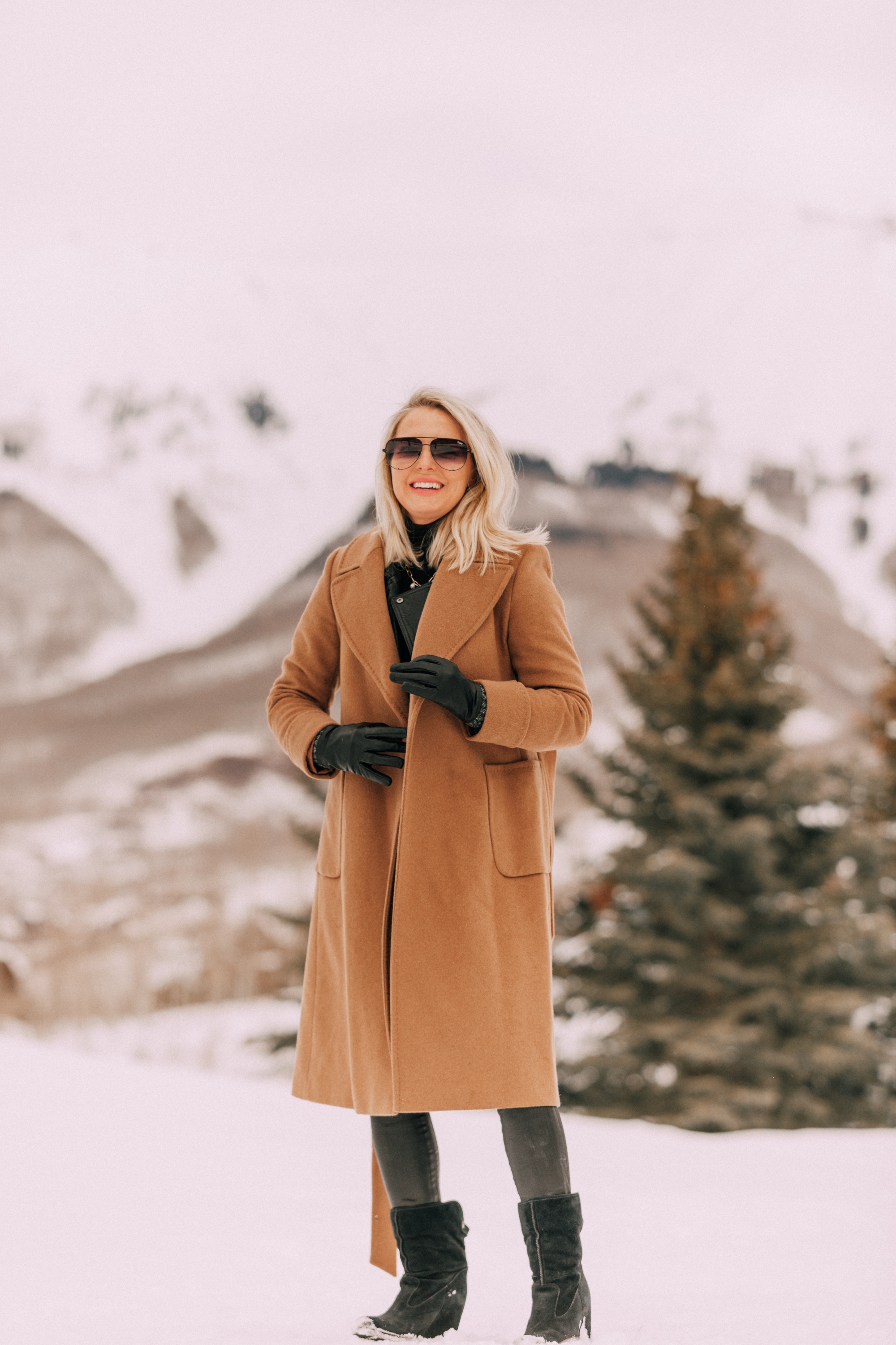 How To Layer A Moto Jacket, Fashion blogger Erin Busbee of BusbeeStyle.com wearing a black leather IRO moto jacket, Carolina Amato gloves, rocket leatherette skinny jeans by Citizens of Humanity, black cashmere turtleneck, and Ugg wedge boots and putting on a wool camel coat by Mackage in Telluride, CO