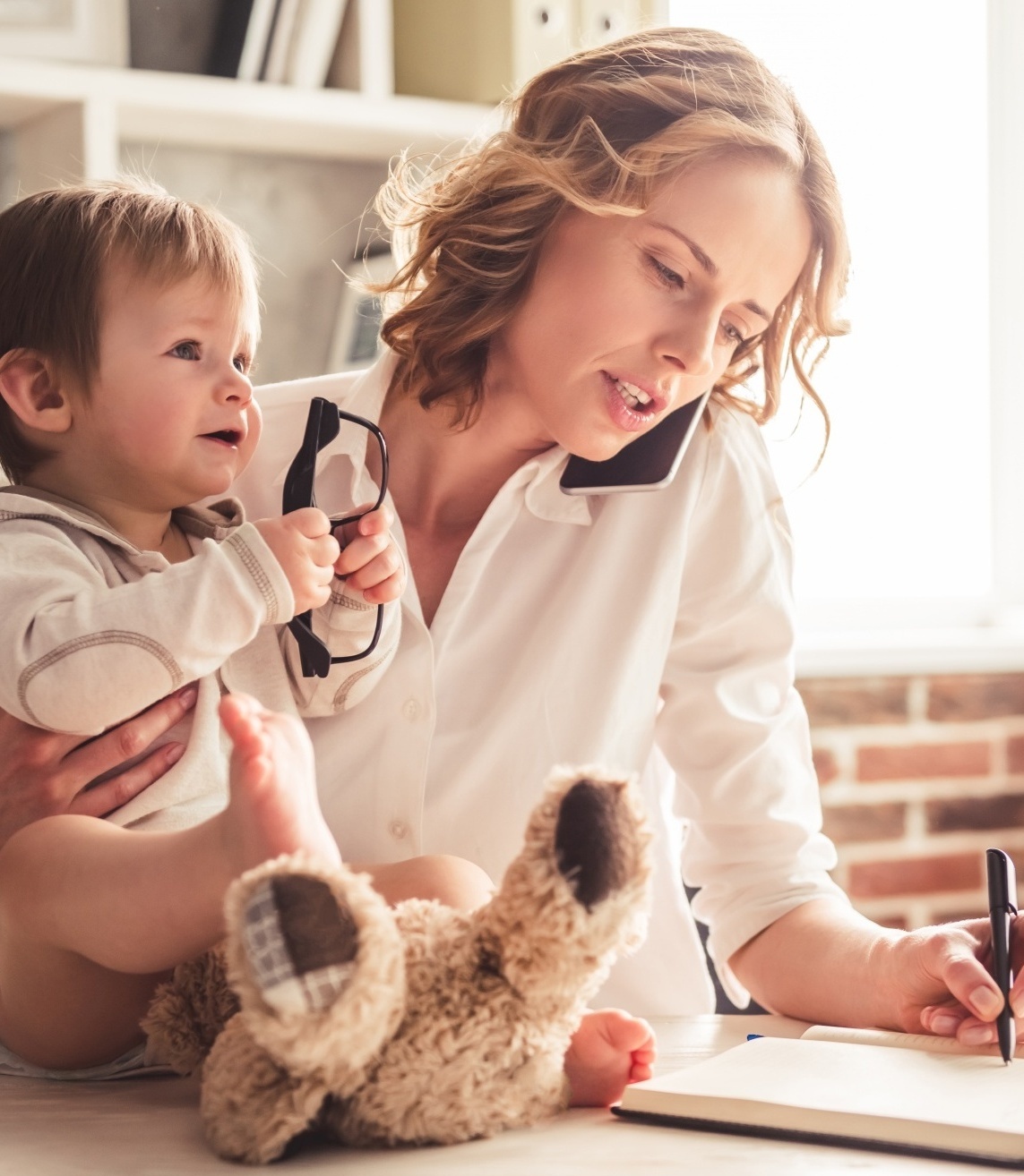 What Saying NO Can Do, blonde woman working and writing holding her baby who has taken glasses and has a stuffed animal