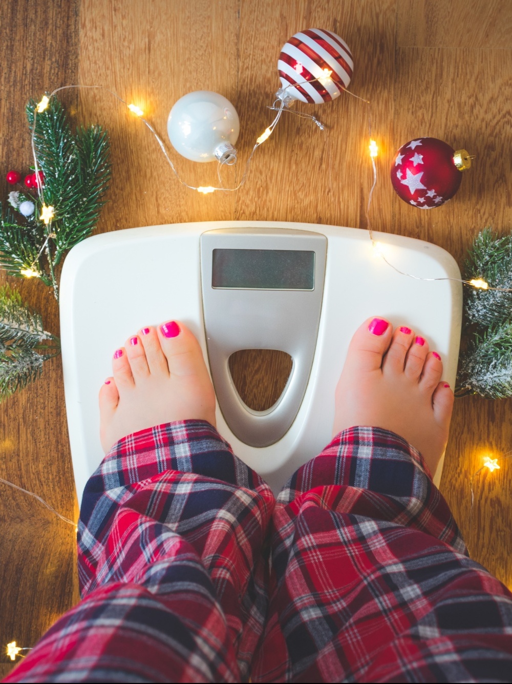 Woman's legs in plaid pj pants standing on bathroom scale surrounded by Christmas ornaments