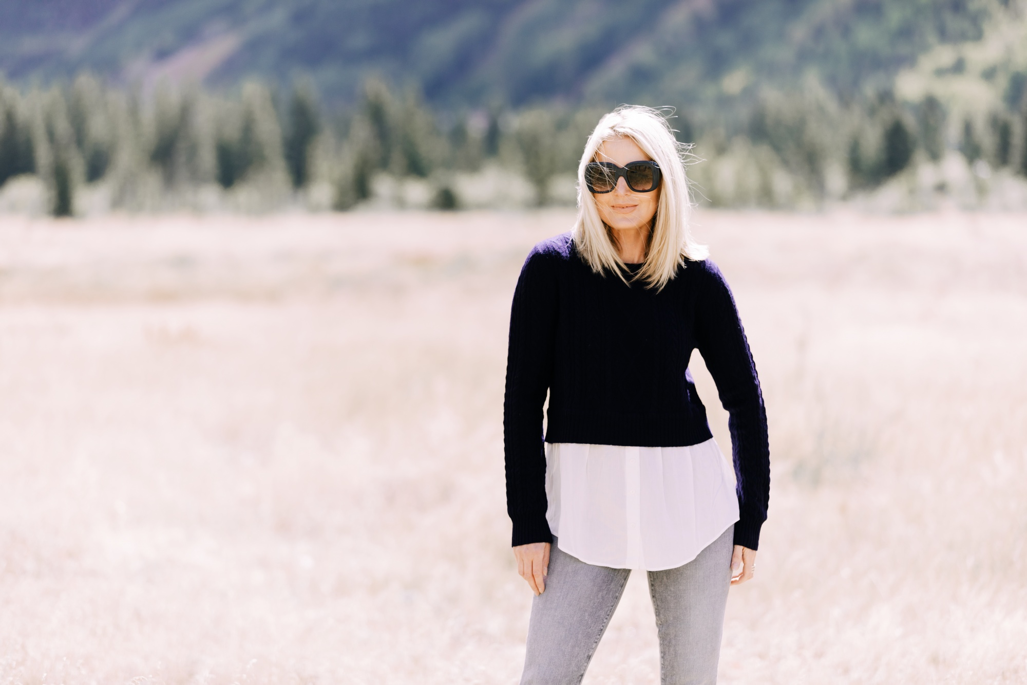 fashion over 40 blogger with blonde hair Erin Busbee in Telluride Colorado with mountains in background wearing navy 2-in-1 sweater with gray skinny jeans and black sunglasses