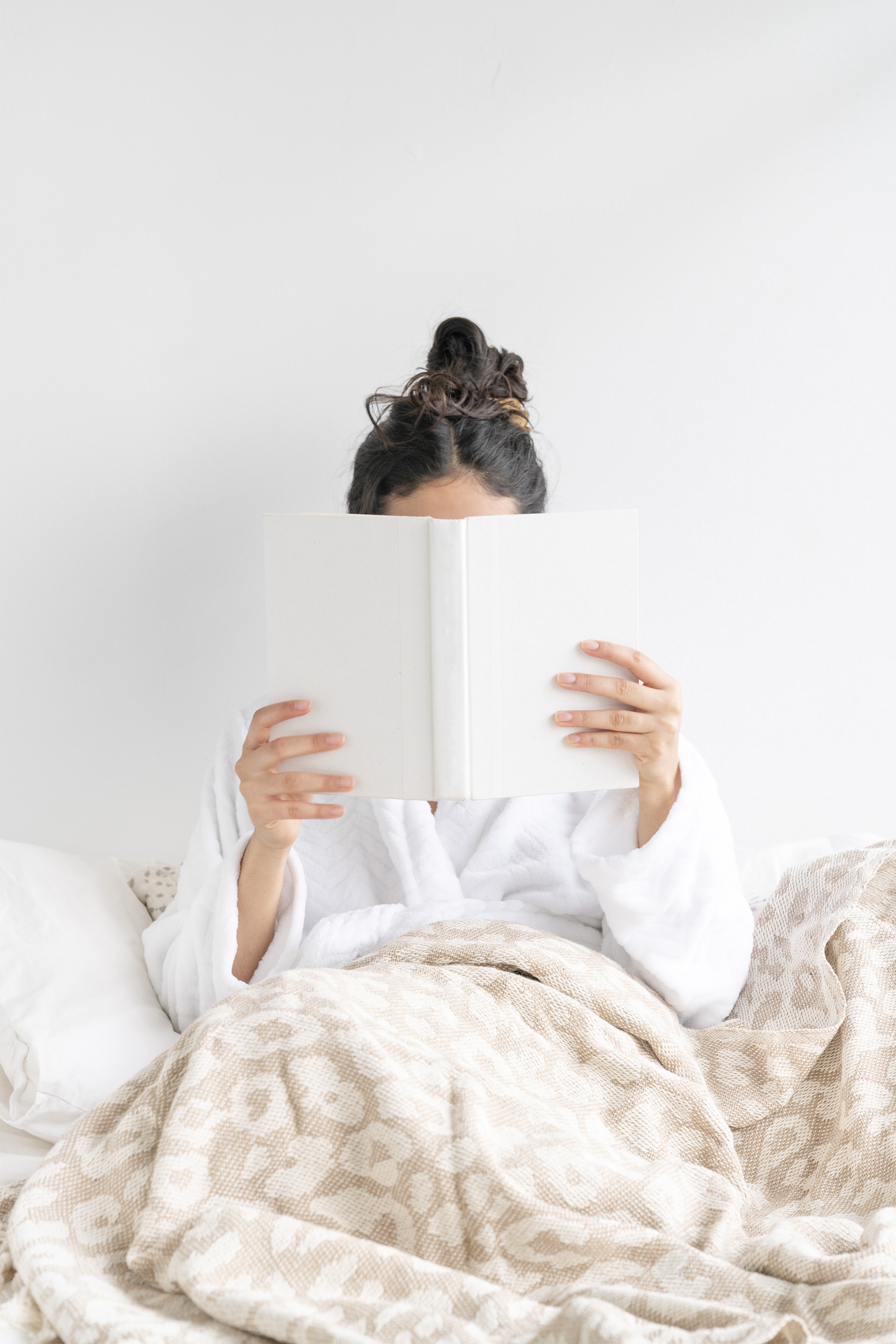 finding hygge at home, cozy-at-home-reading-a-book-in-bed-wtih-soft-neutral-colored-leopard-blanket-wearing-a-white-robe.