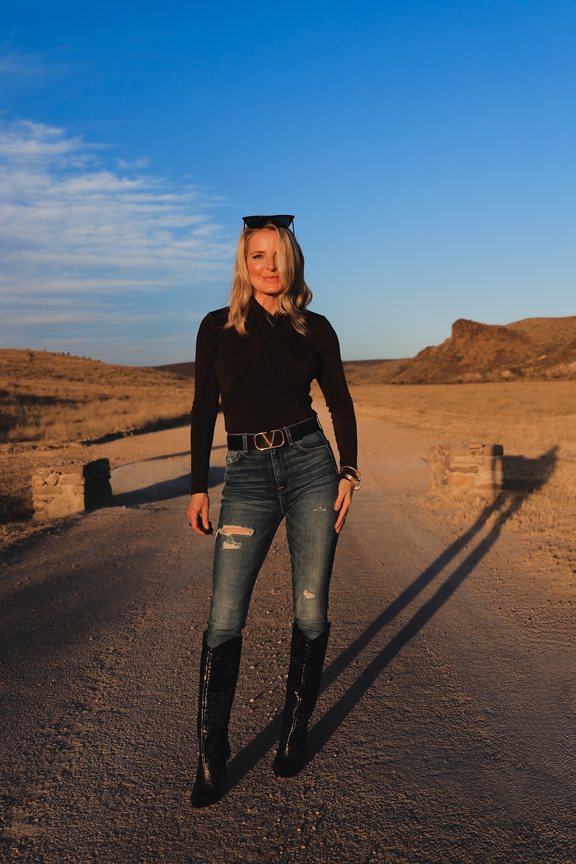 Brown and black outfit, Erin Busbee of Busbee Style wearing a dark brown wrapped neck top by Lovers + Friends tucked into 7 for all mankind ripped jeans with black snake-embossed knee-high boots and black reversible Valentino belt in west Texas