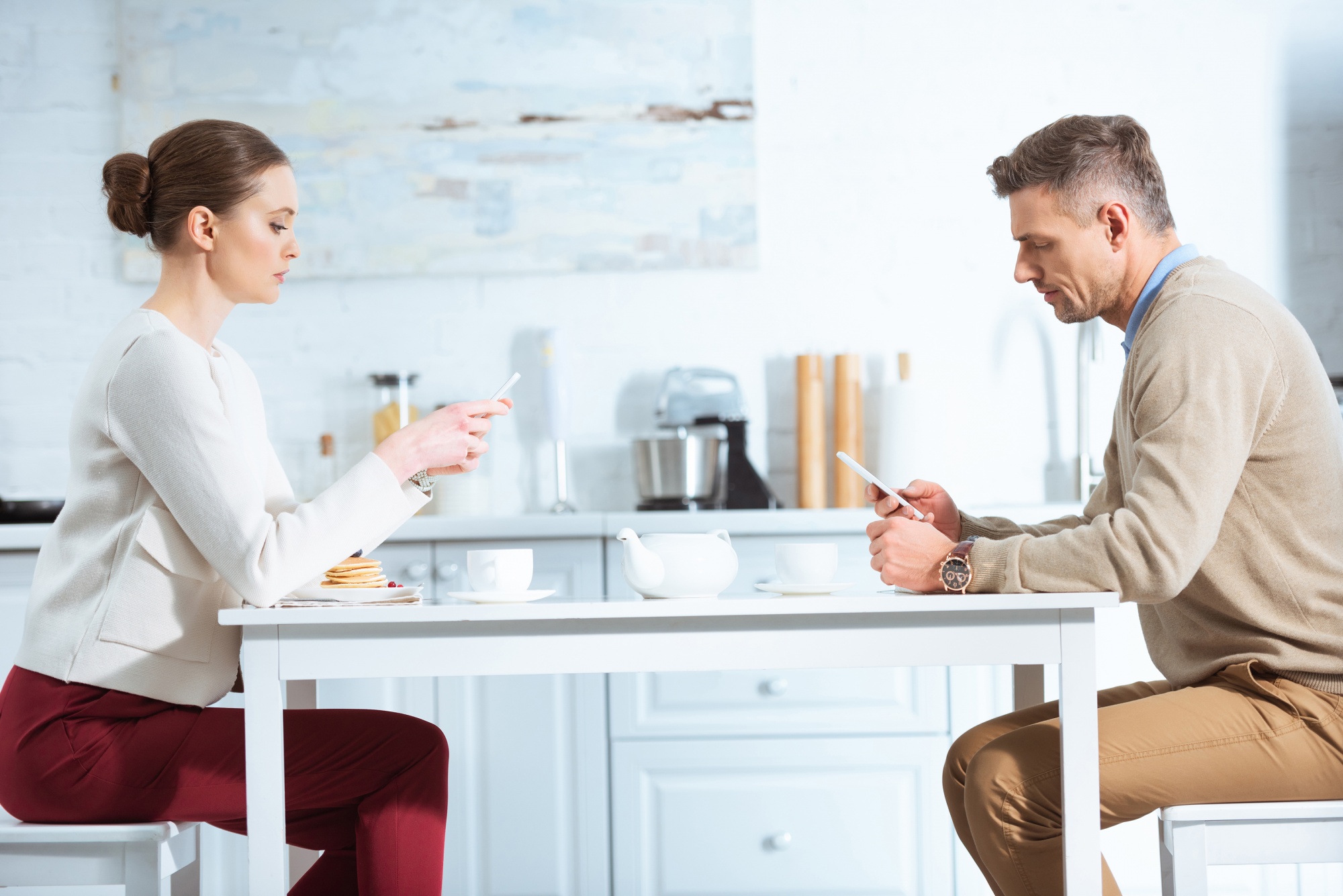 Toxic relationship habits, couple sitting at kitchen table ignoring each other and focusing on their cell phones