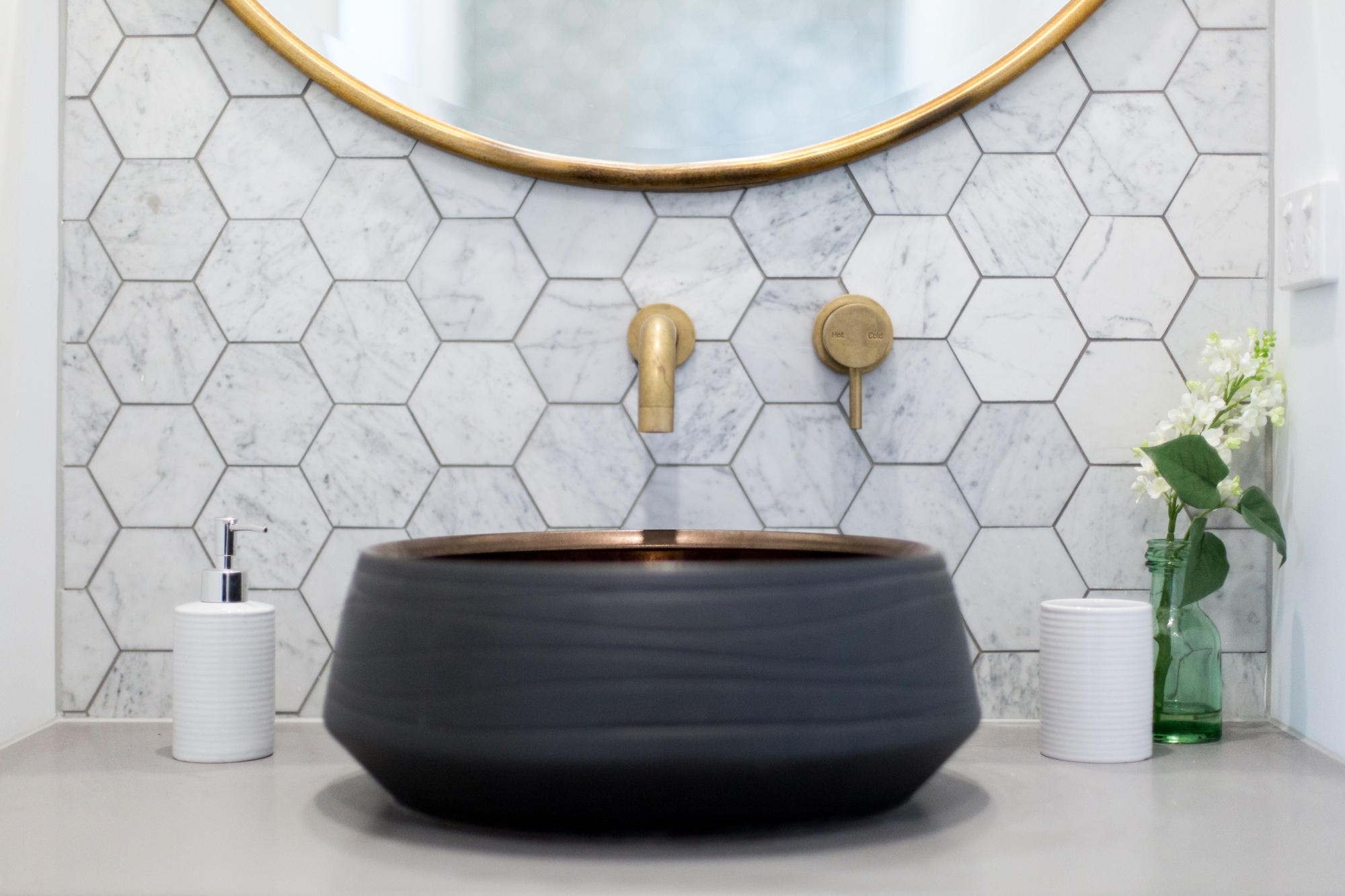 How To Update Your Powder Room With These 4 Easy Changes featuring an updated vanity with a large, round, brass mirror, small vase of flowers and white soap dispenser