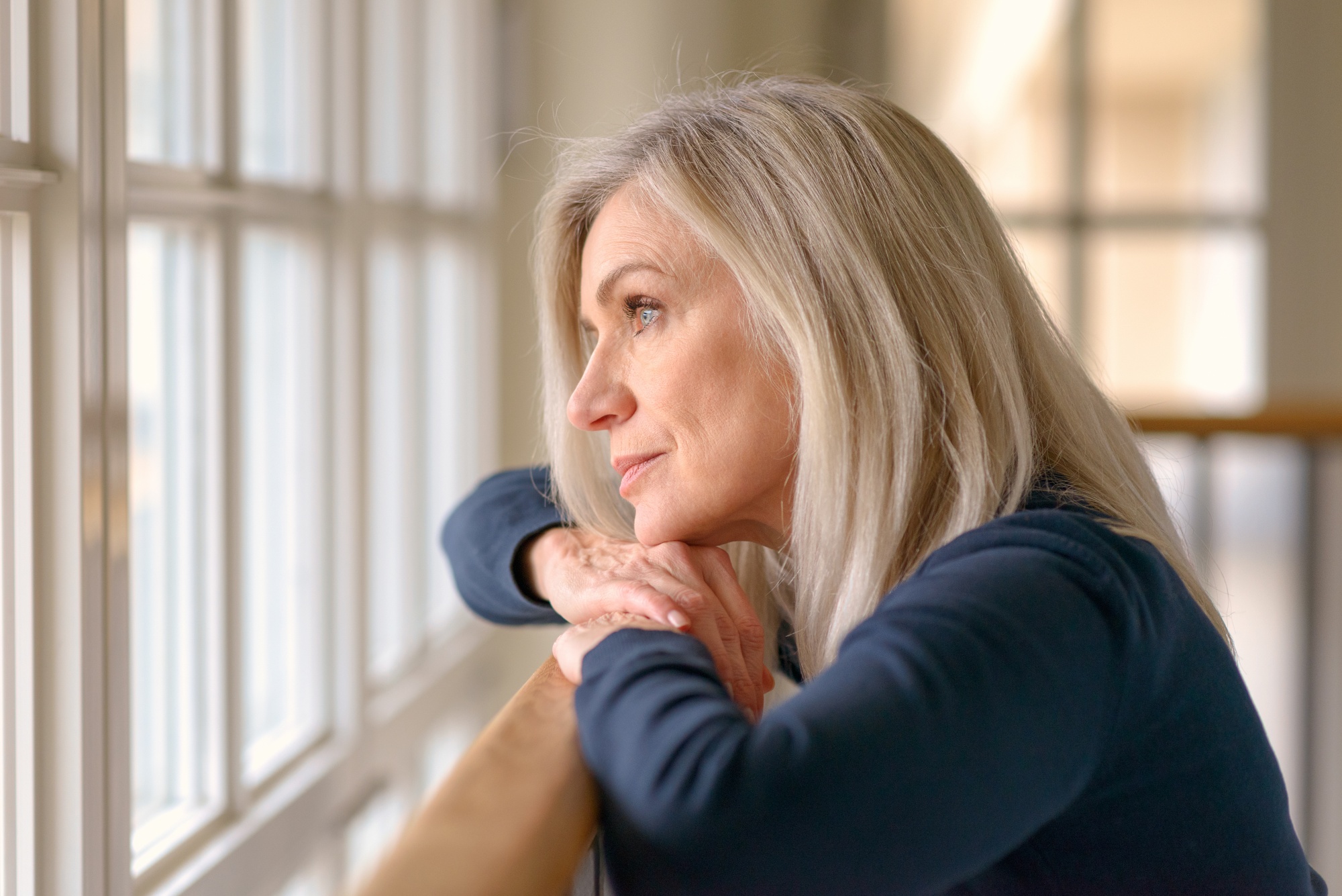 Embrace negative thoughts and turn them into joy, Middle-aged blonde woman in blue shirt looking out window a little sad