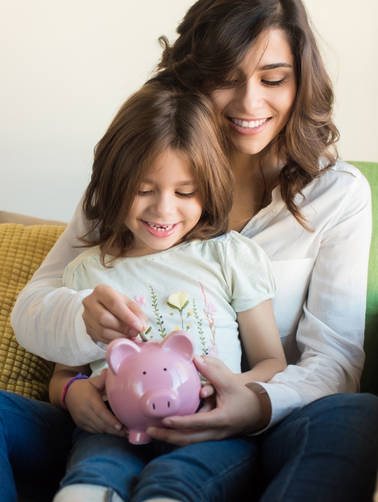 Teaching kids about money, mom and daughter sitting on the sofa putting coins in a piggy bank