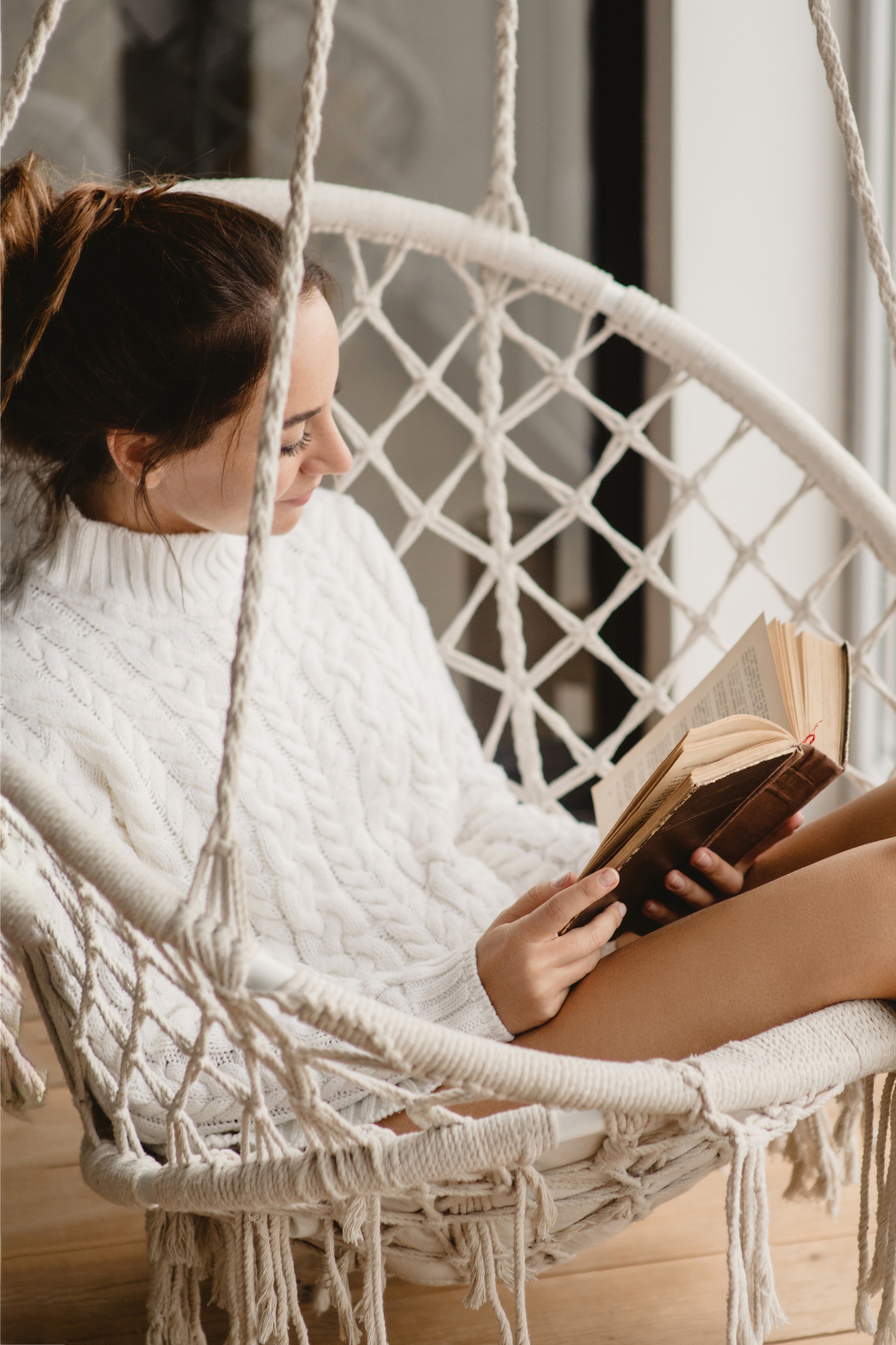 self-care and self-love, woman in cableknit white sweater reading in hammock swing
