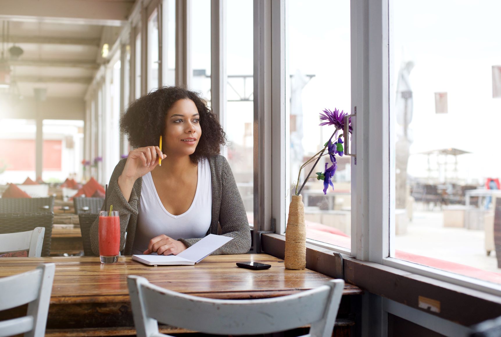 Black woman in white V-neck and grey cardigan at diner table writing and looking out window, manifestation for beginners, manifest your dream life