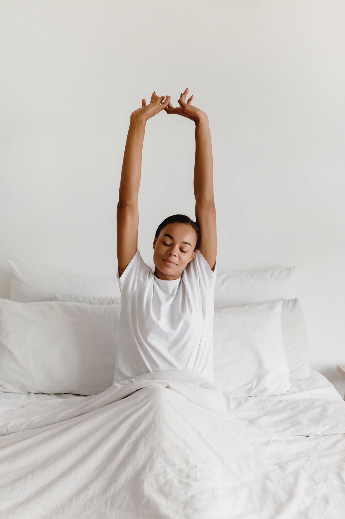 Black woman in white pjs in white bed with arms overhead stretching - how to get better sleep, sleep routines, sleep better tonight