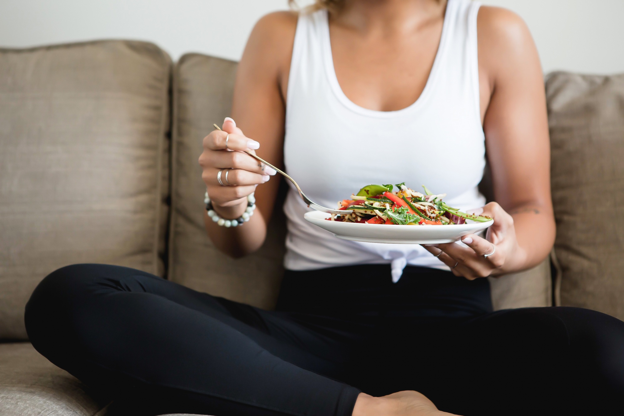 intermittent fasting and intuitive fasting, Woman in white tank and black leggings eating salad on her couch